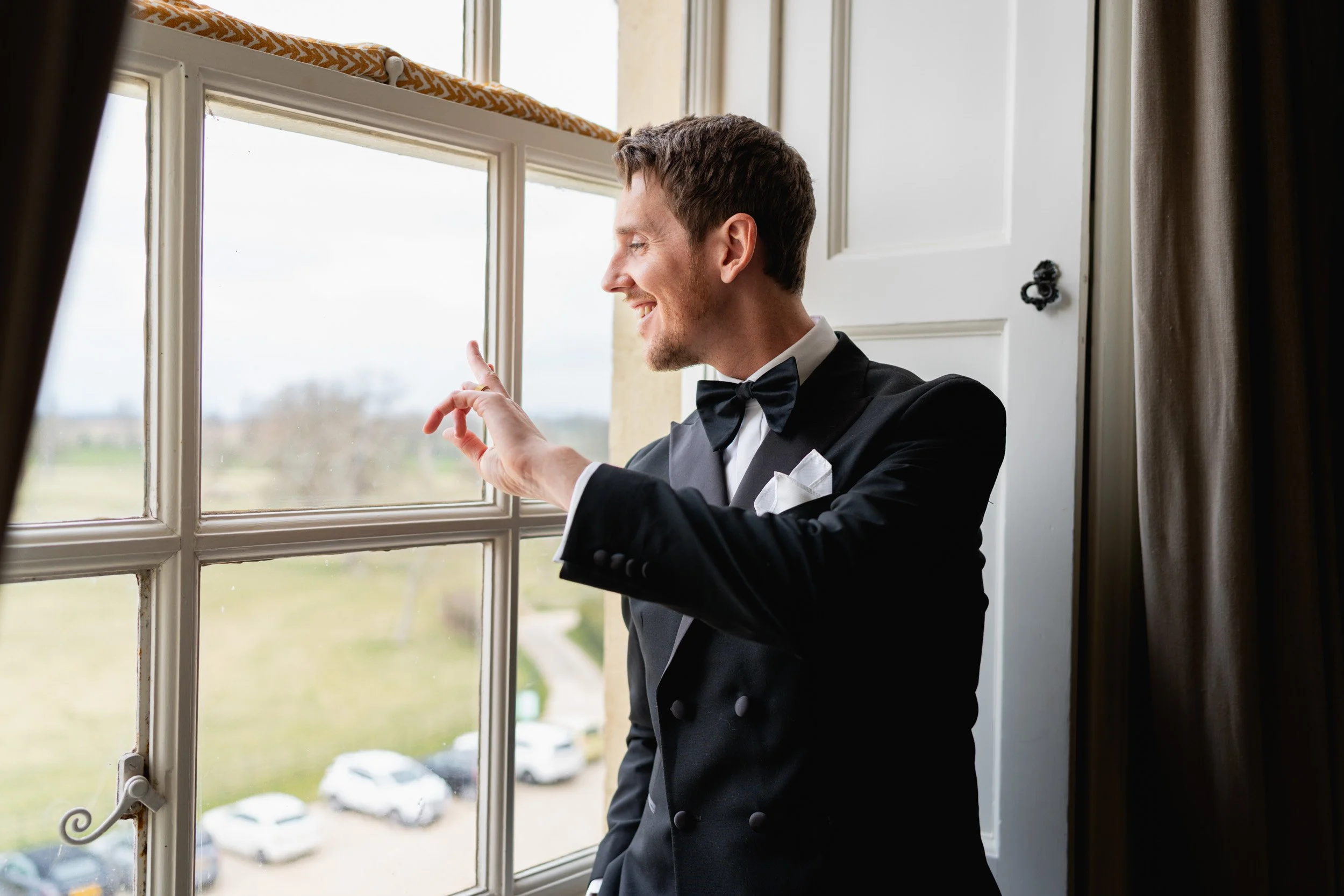 Groom waving from a bedroom window as wedding guests arrive at Kirtlington Park in Oxfordshire