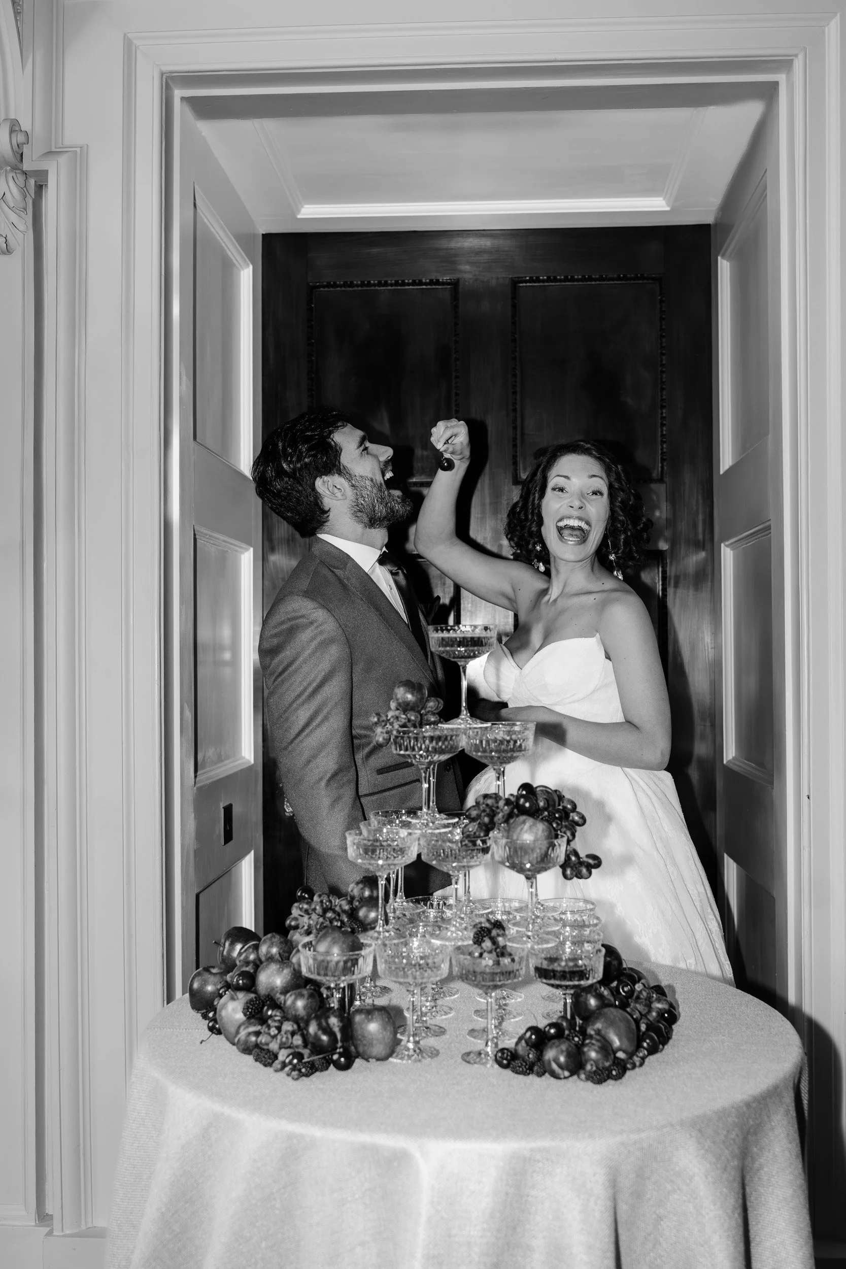 Bride cheerfully feeding her groom a cherry beside the champagne tower at their reception  at Kirtlington Park in Oxfordshire
