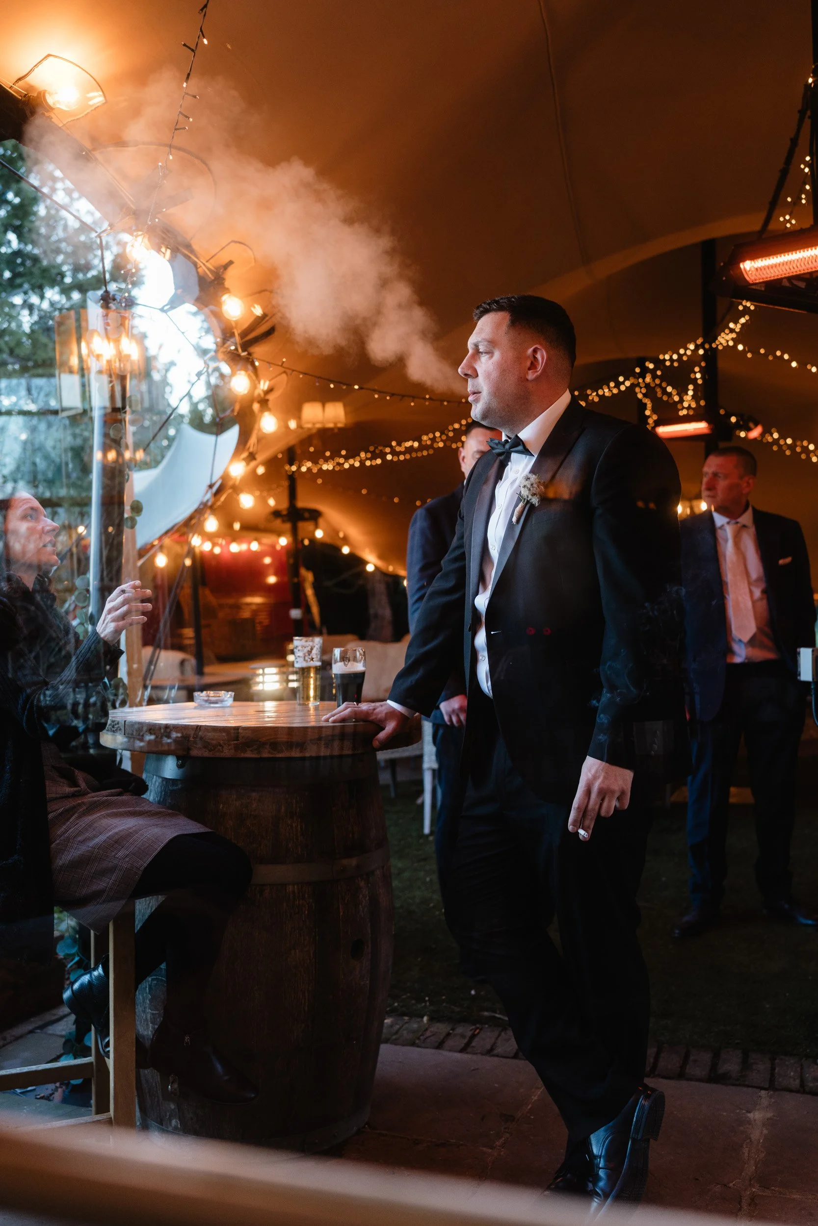 Wedding guest smoking a cigarette in the softly lit marquee during an evening reception at The Bedford Arms in Chenies, Buckinghamshire