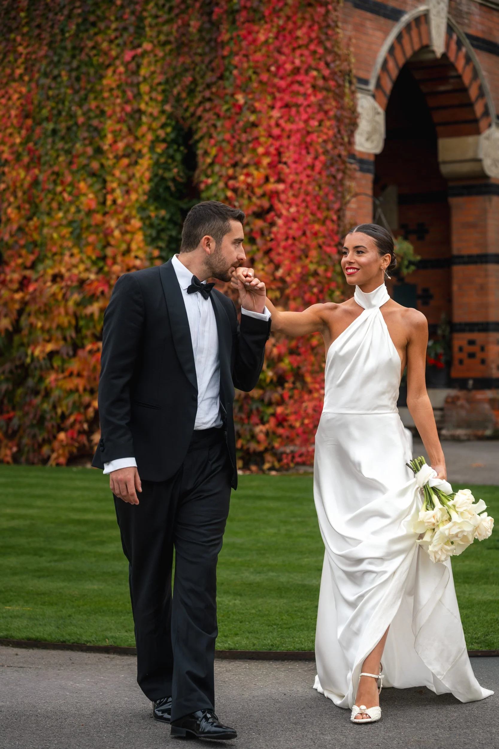 Groom leading his bride whilst kissing her hand outside The Elvetham Hotel in Hampshire with ivy-covered red brick architecture behind them 