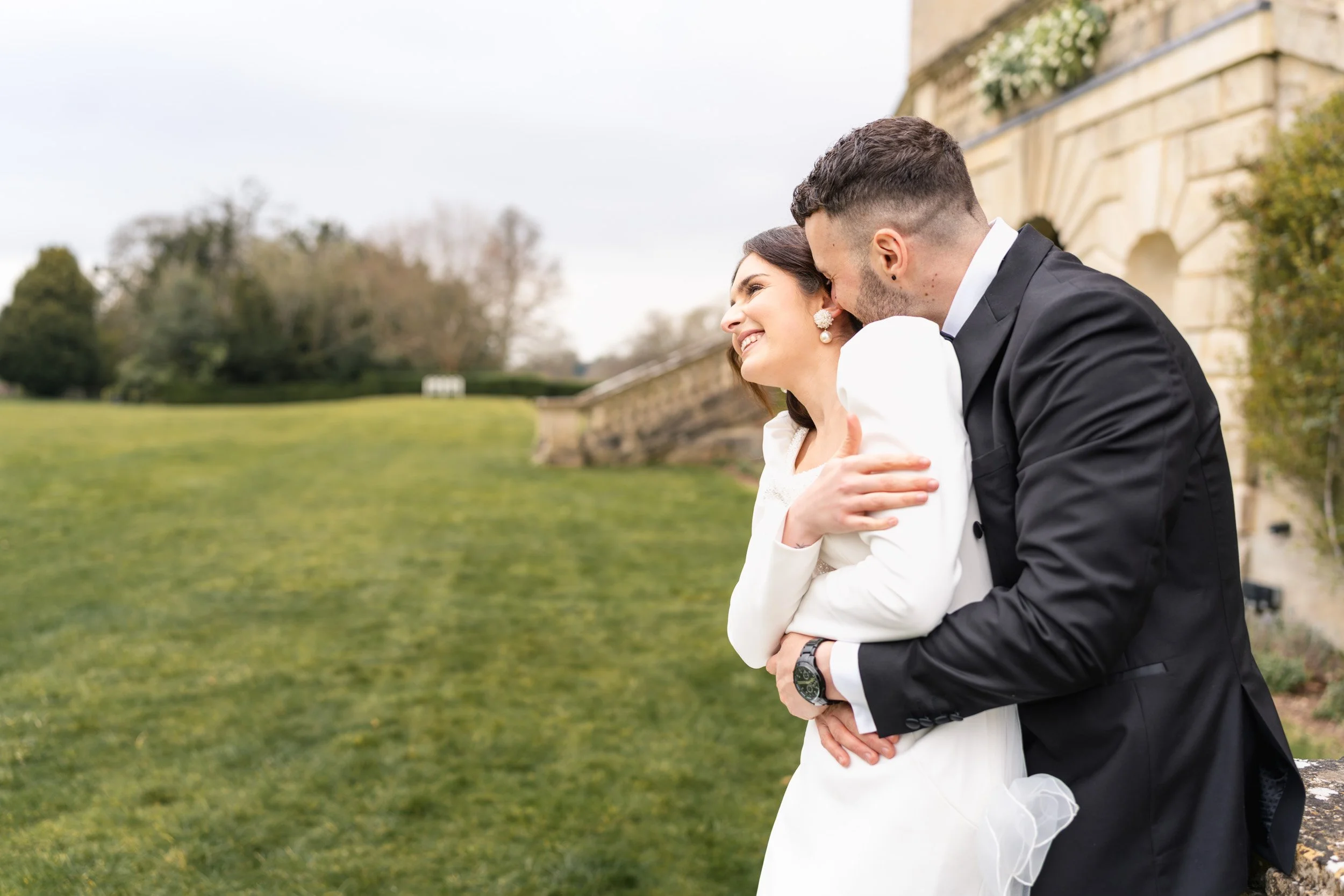 Groom pulling bride close and whispering in the ear of his bride beside the balustrade in the gardens at Kirtlington Park in Oxfordshire