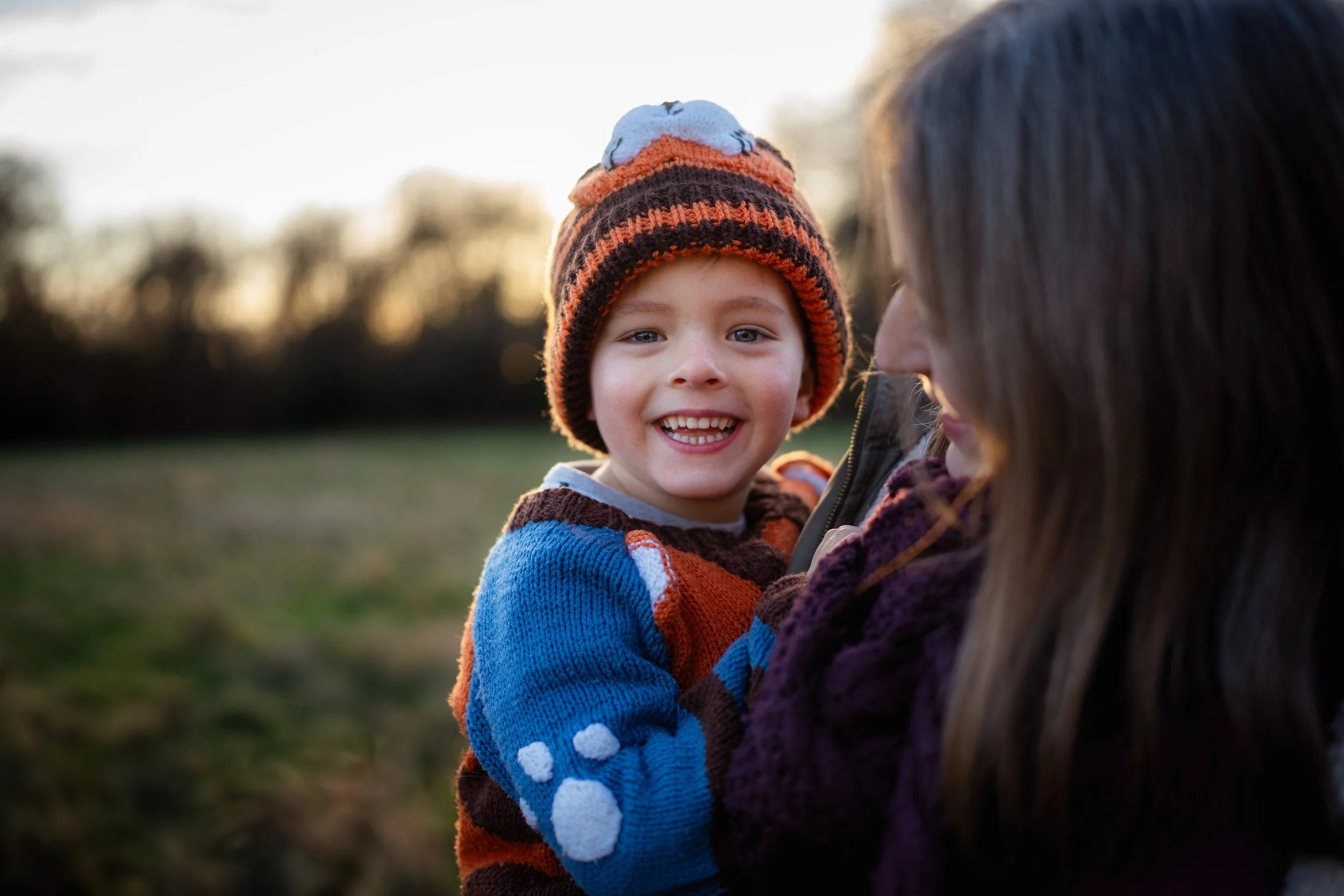 A mother smiles at her happy boy wearing a bobble hat in a relaxed outdoor family photography session in Amersham, Buckinghamshire a golden hour
