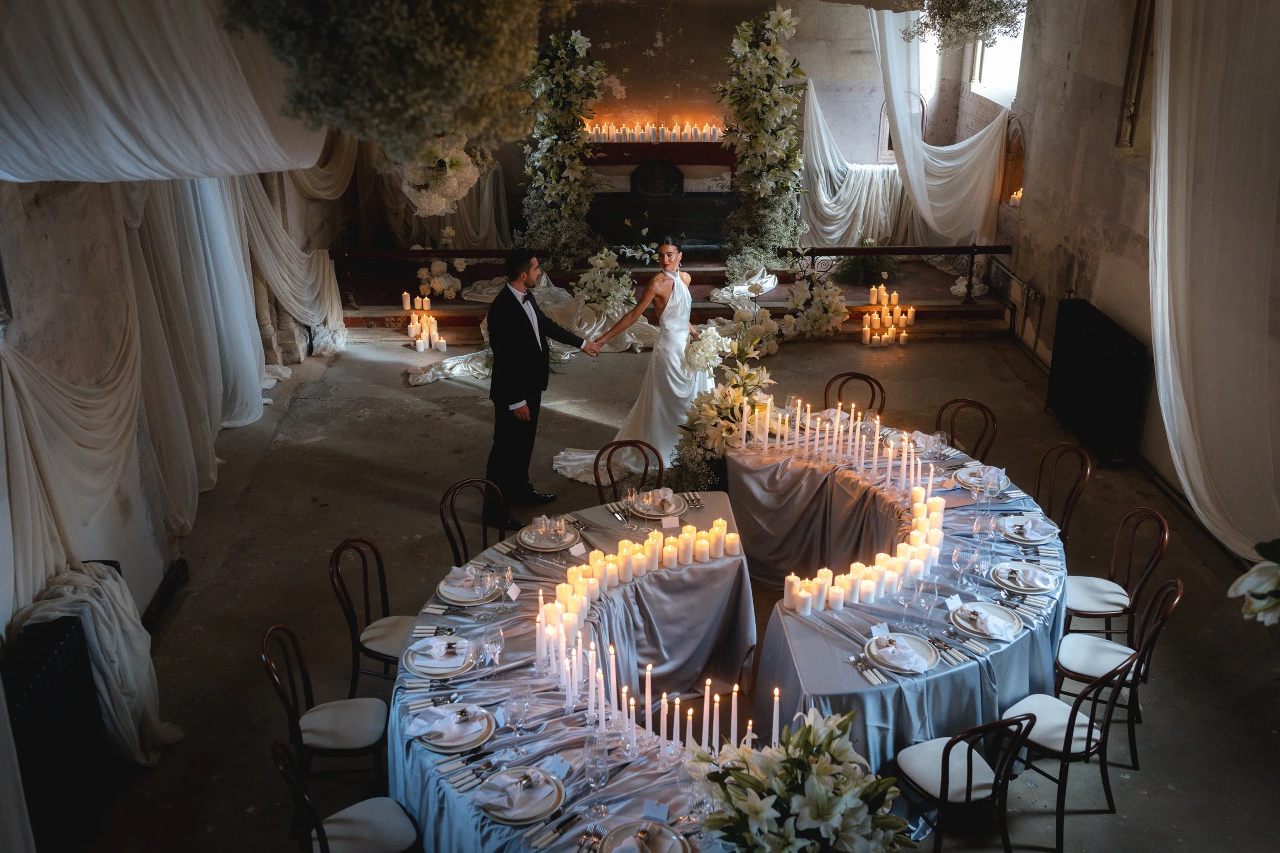 Bride and groom walking head table lit by candle light and covered in silks and white florals wedding reception inside The Bell Tower at The Elvetham Hotel in Hampshire