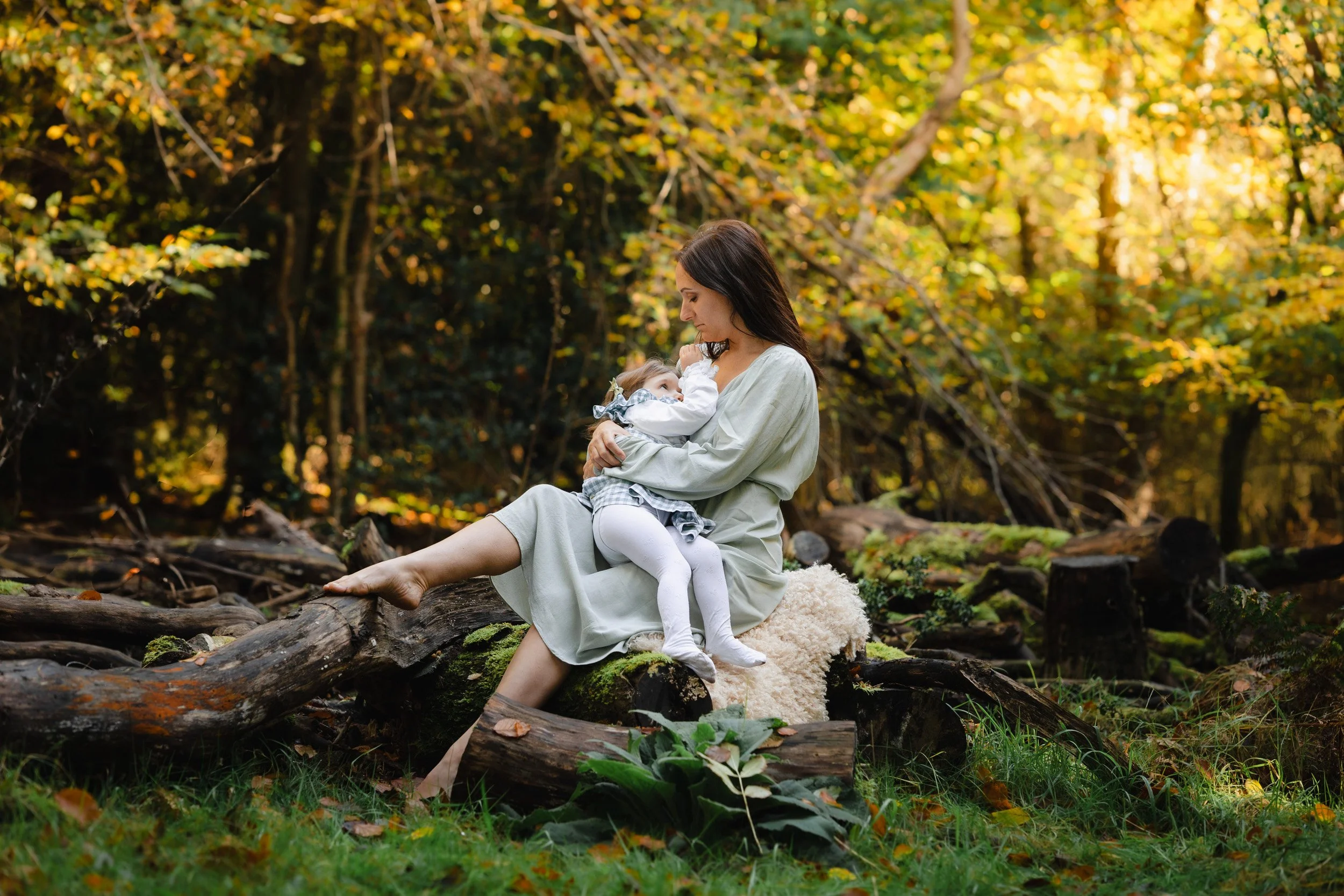 A mother sitting on a log in the golden light of a woods in Amersham, Buckinghamshire feeding her baby 
