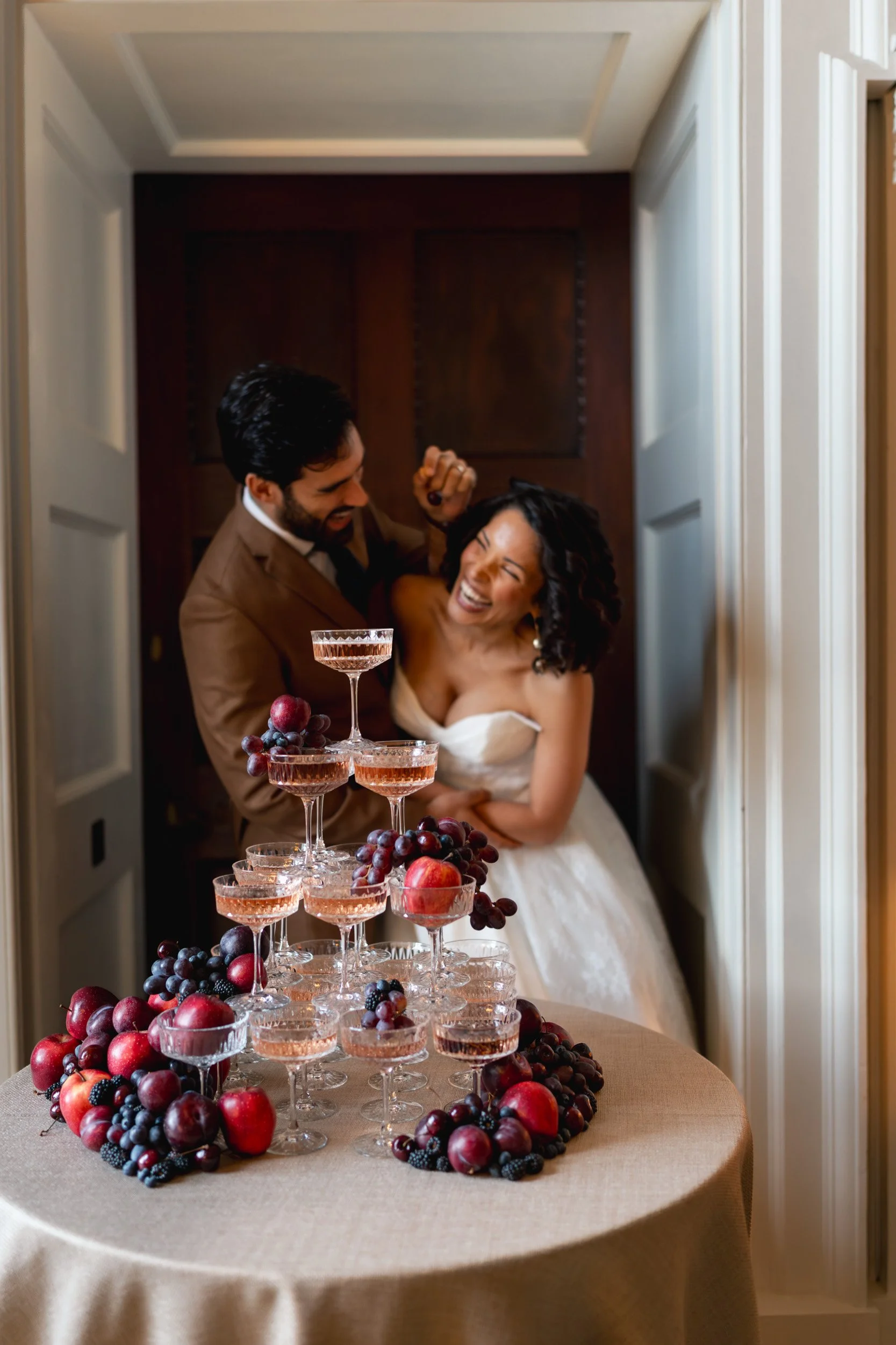 groom cheerfully feeding his bride a cherry beside the champagne tower at their reception  at Kirtlington Park in Oxfordshire