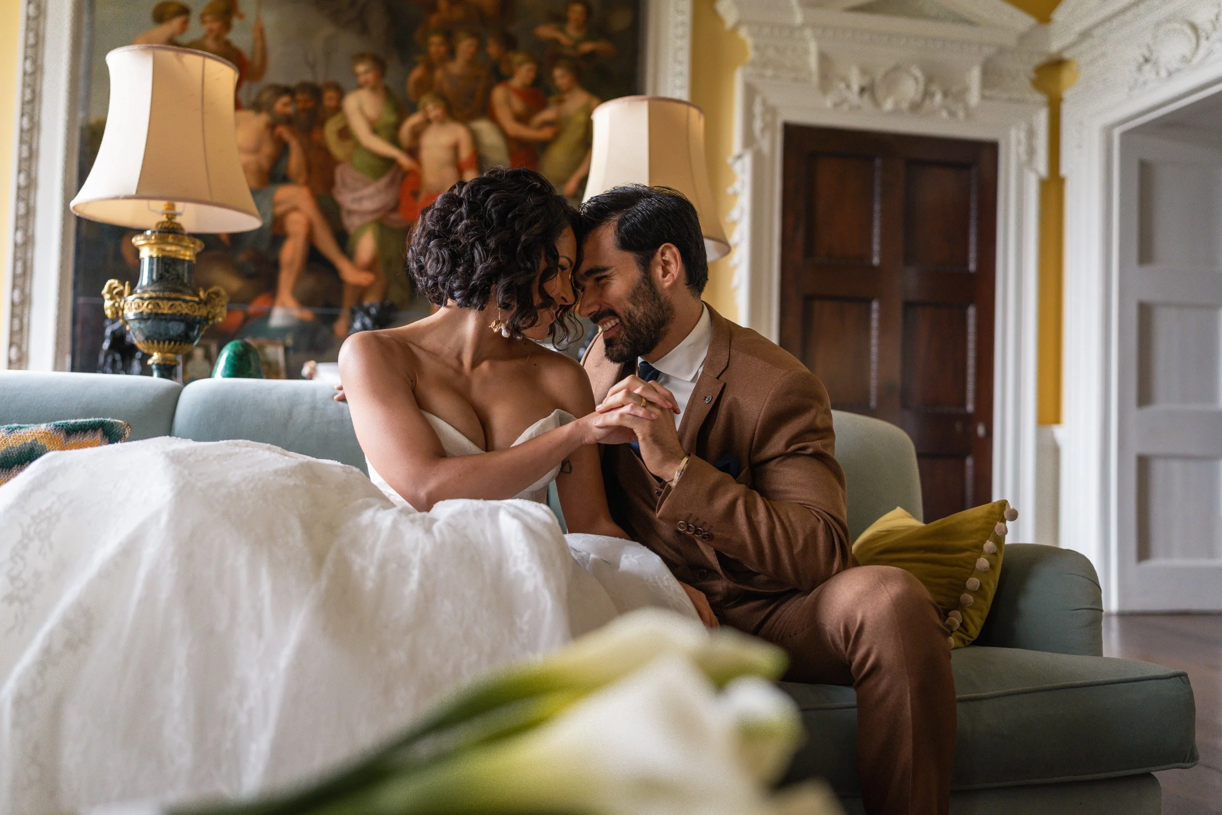 Bride and groom sitting close together on a sofa in an elegant room at Kirtlington Park in Oxfordshire