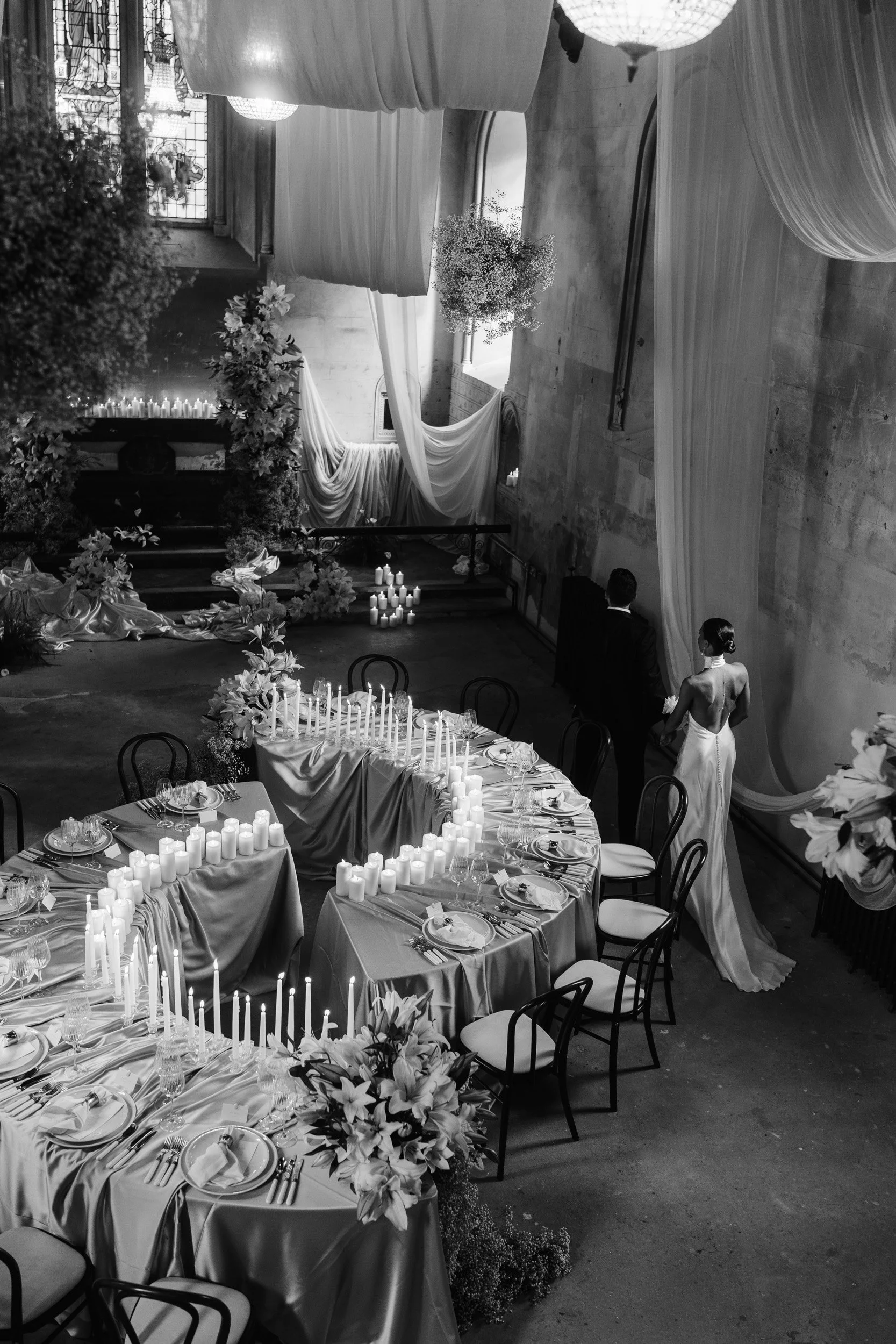 Bride and groom walking through a candlelit reception room with draped fabric and floral displays inside The Bell Tower at The Elvetham Hotel in Hampshire