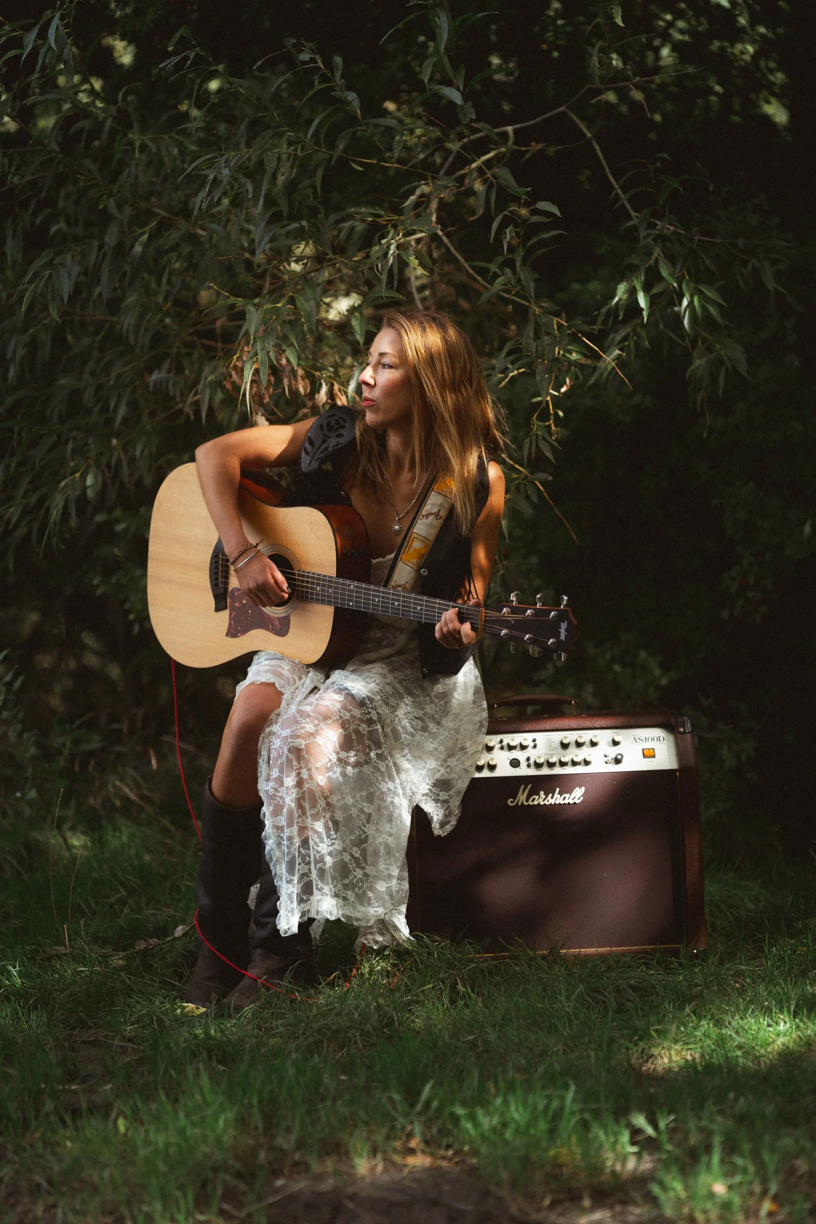 Woman musician playing her acoustic guitar in a lace dress sat on her amp for a personal branding portrait by the River Thames in Windsor Berkshire