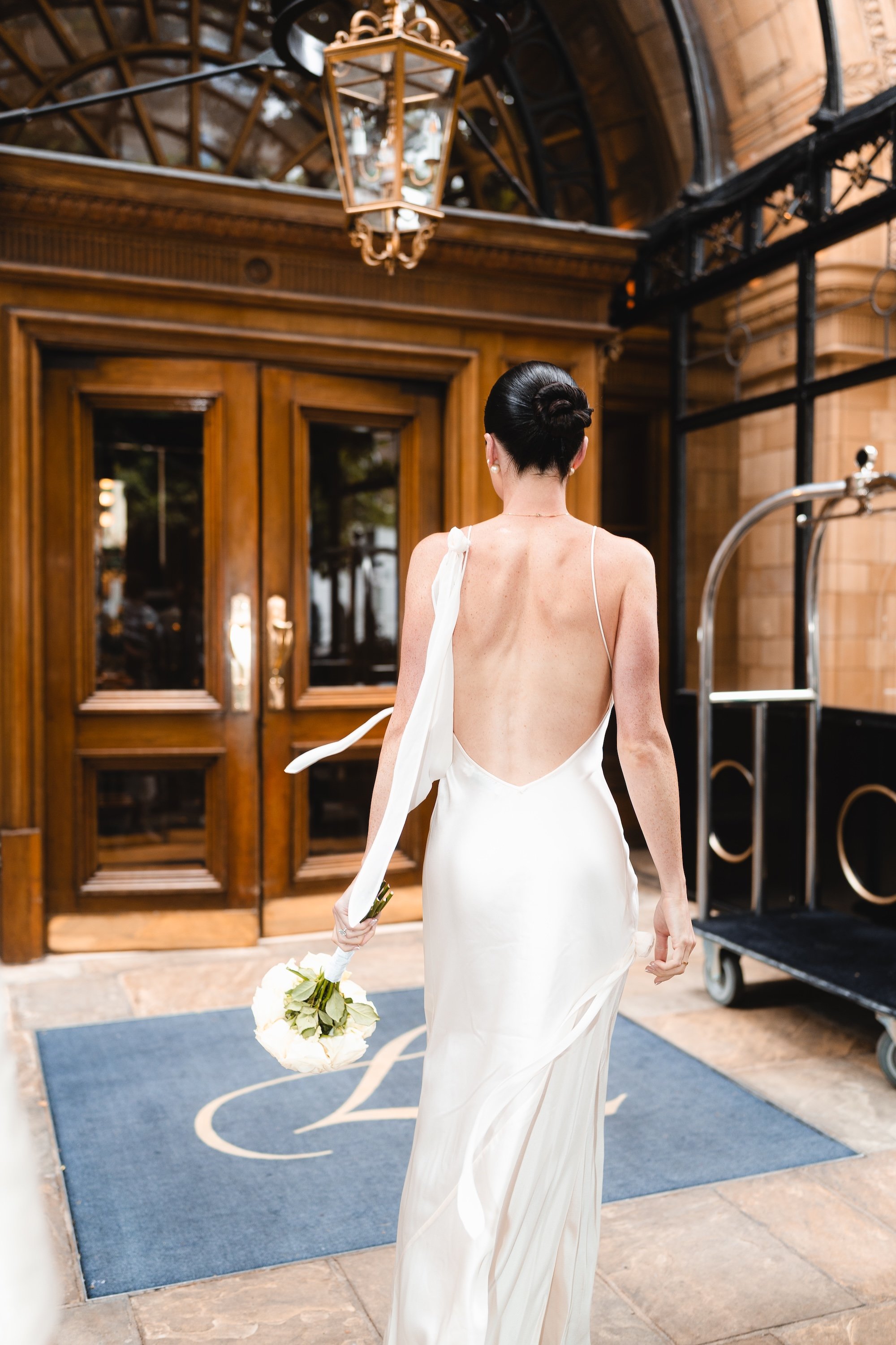 Bride in a beautiful backless wedding gown holding her flowers walks into The Landmark London after saying goodbye to her wedding photographer after her Marylebone Town Hall civil wedding ceremony
