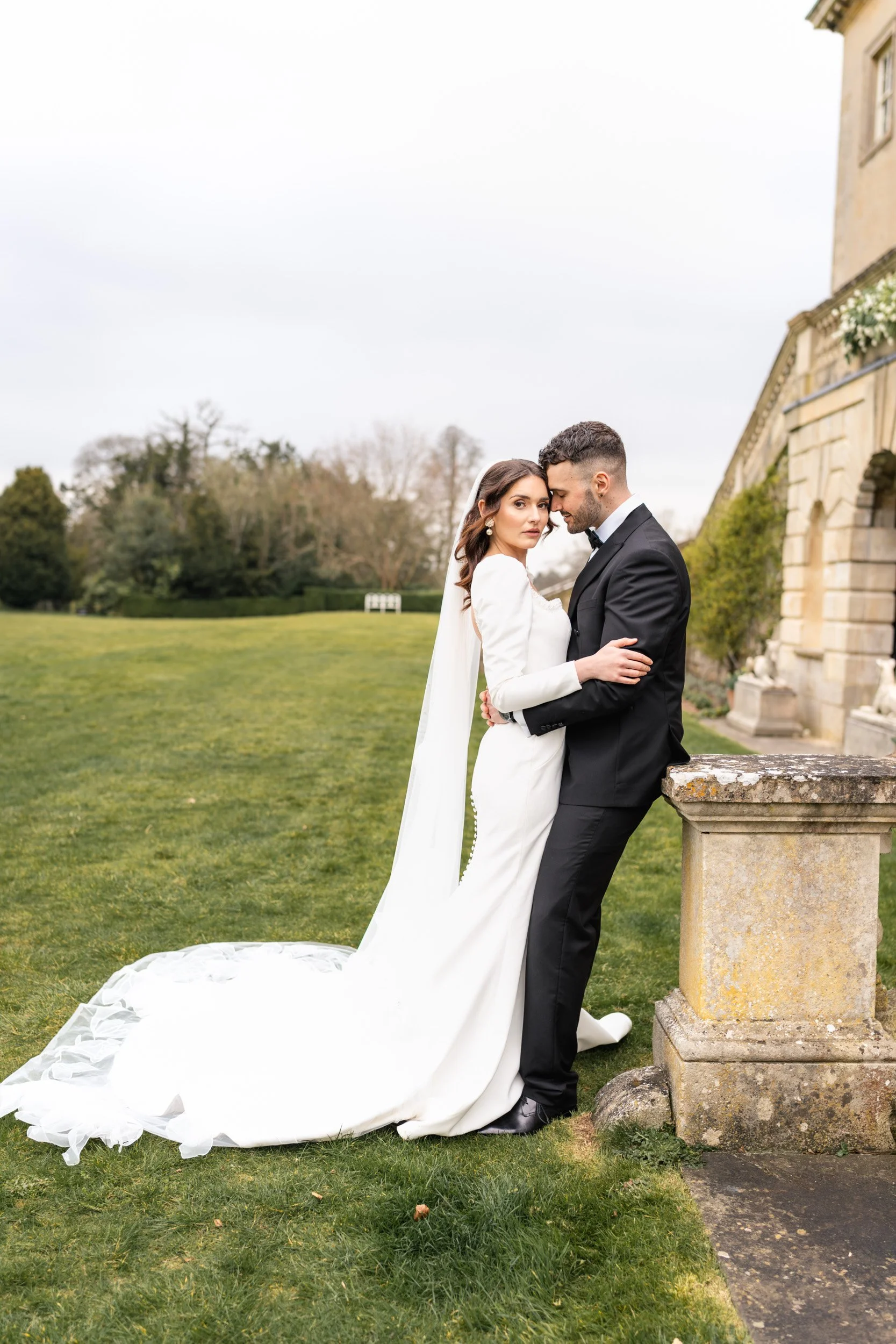 Newly wed Bride and groom cuddle by the stone steps on the grounds of Kirtlington Park in Oxfordshire just before sunset