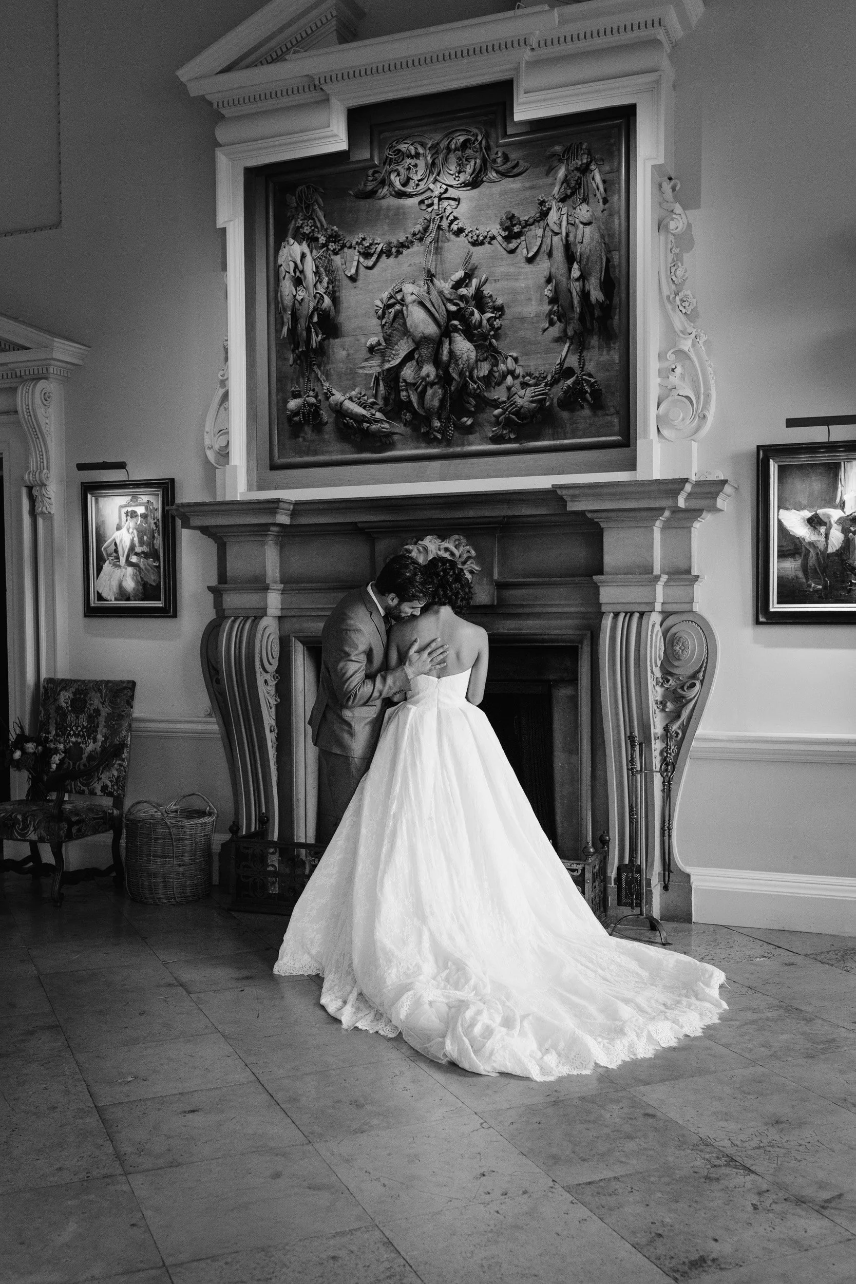 Bride and groom having an intimate moment as she leans in to him and he kisses her on the shoulder in front of the fire place at their wedding reception at Kirtlington Park in Oxfordshire