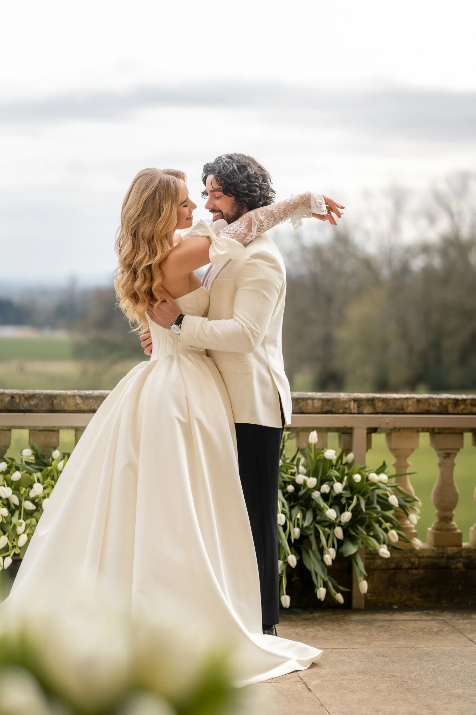Bride and groom embracing on the white tulip lined balcony over looking the grounds and gardens of Kirtlington Park in Oxforshire