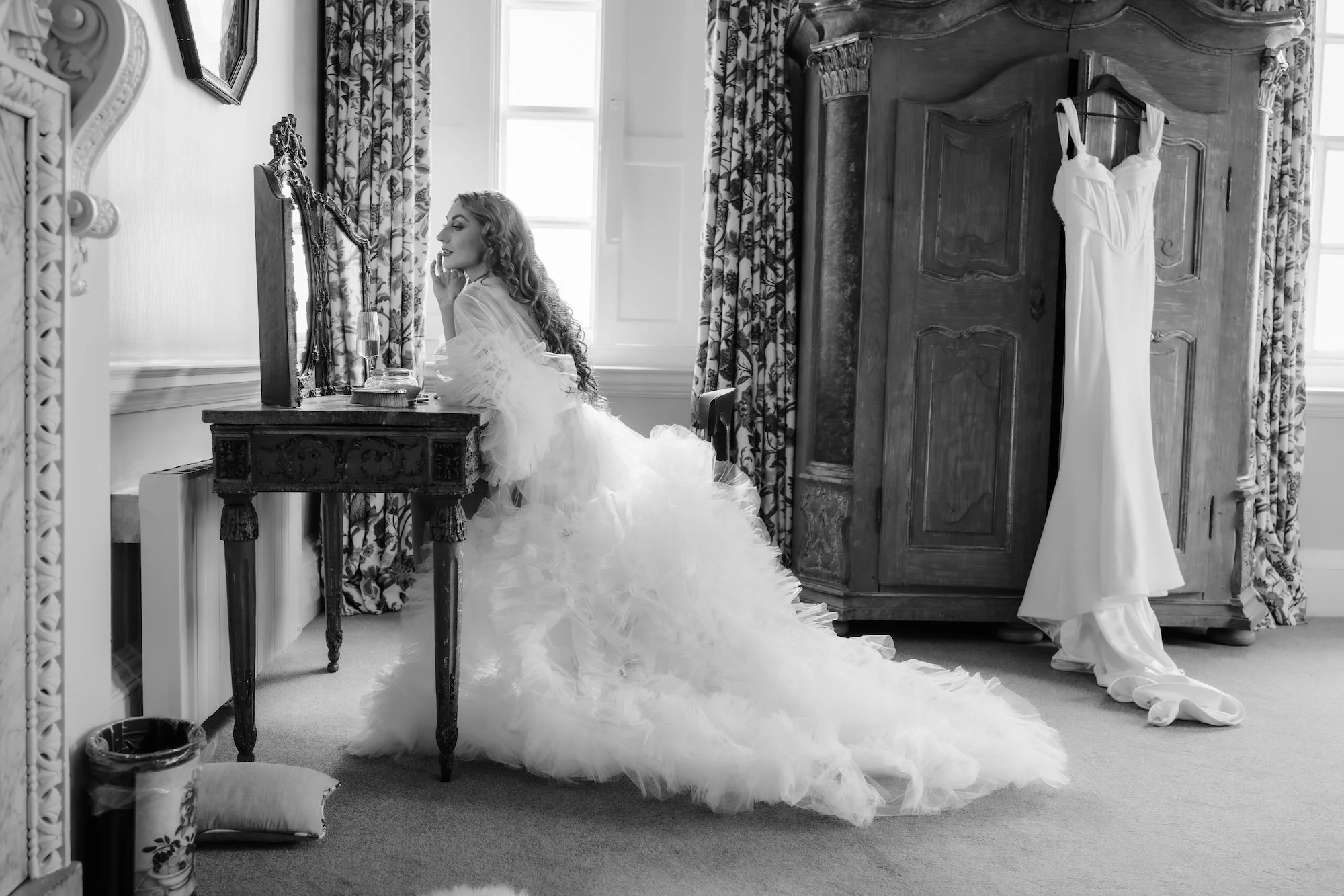 Bride at her dressing table in the Bridal suite of Kirtlington Park. Wedding dress hangs on the antique wardrobe