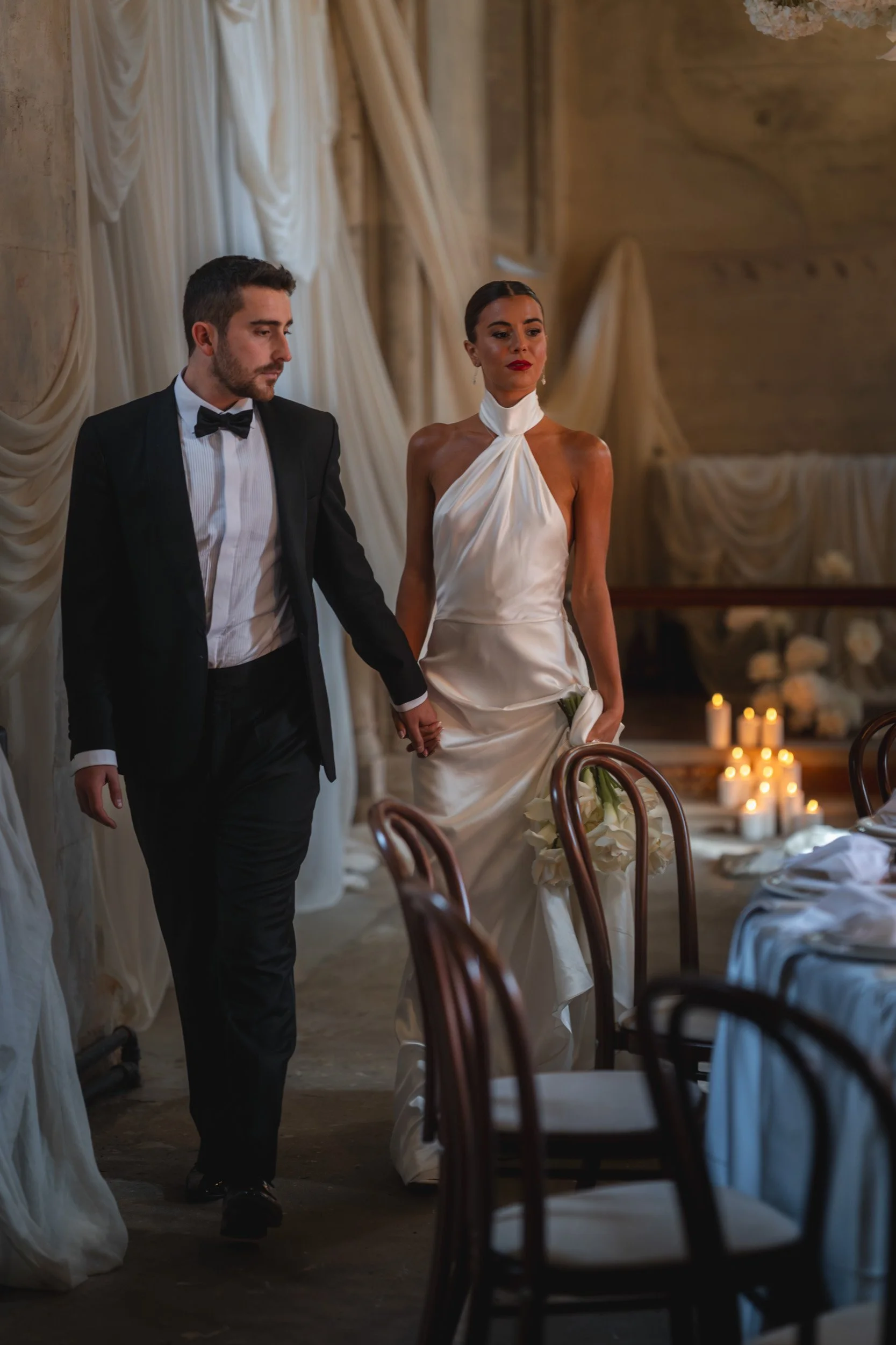 Bride and groom walking through a candlelit reception room with draped fabric and floral displays inside The Bell Tower at The Elvetham Hotel in Hampshire