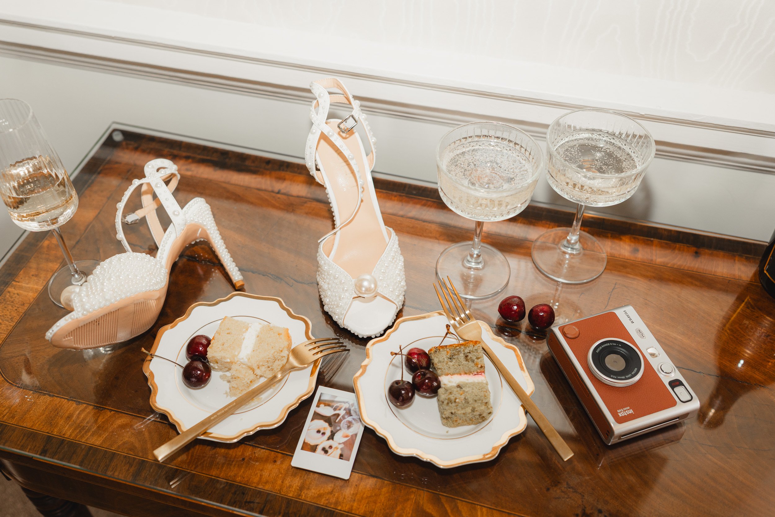 Wedding details including bridal shoes, champagne glasses, cake and instant camera in a suite at Kirtlington Park in Oxfordshire