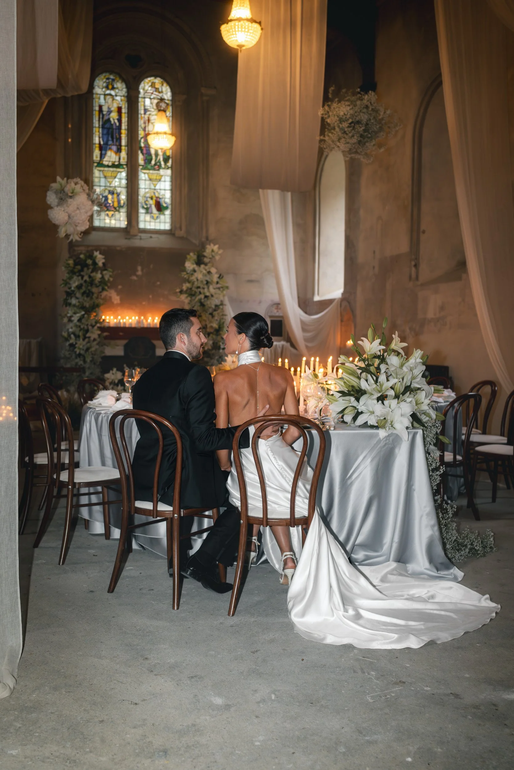 The bride and groom sitting close together in their  candlelit reception room with flowing drapery and long banquet tables inside The Bell Tower at The Elvetham Hotel in Hampshire