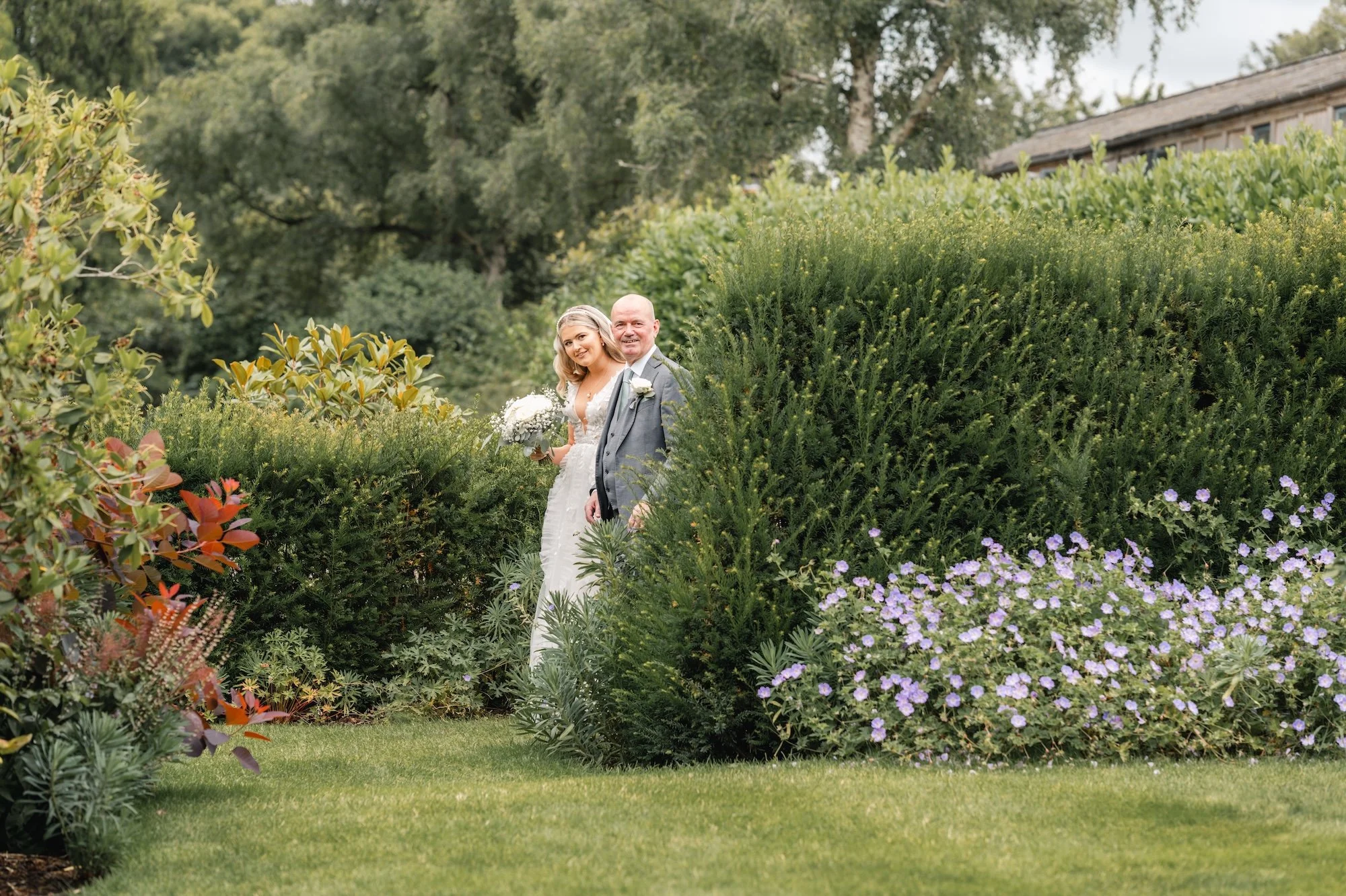 Father of the bride walking his daughter towards her outdoor wedding ceremony at Wasing Park, glimpsed through the garden hedges