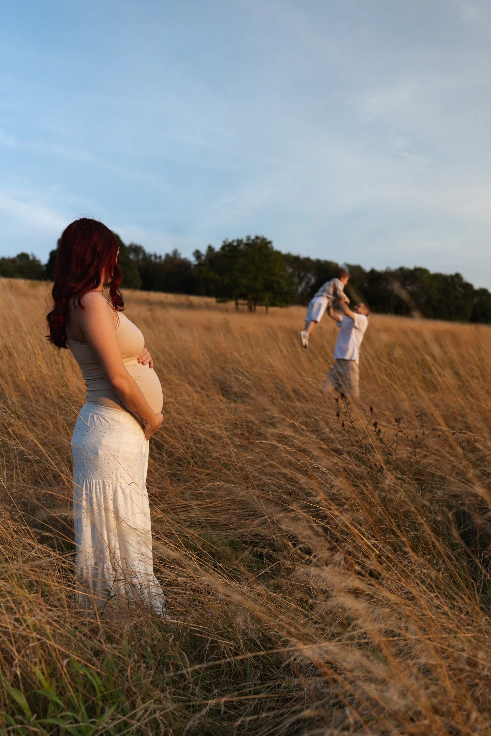 a pregnant mother holds her belly in the golden sunlight of a field in Great Missenden in Buckinghamshire looking on at her husband playing with their toddler