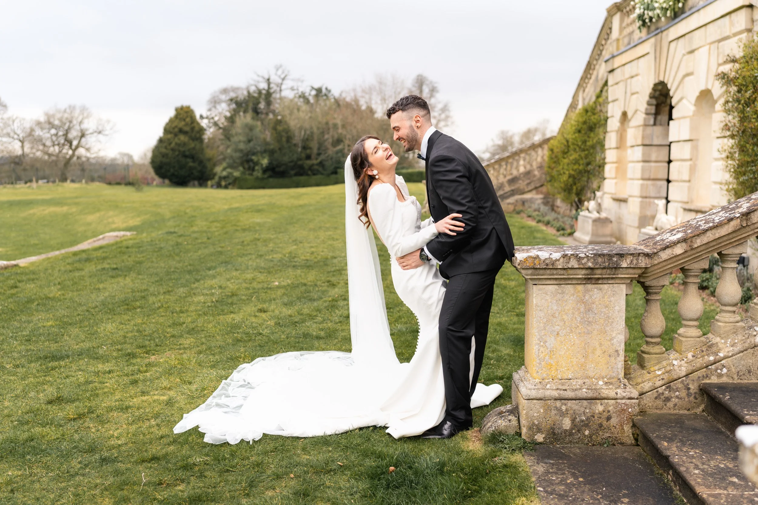 Bride and groom laughing and hugging beside the balustrade in the gardens at Kirtlington Park in Oxfordshire