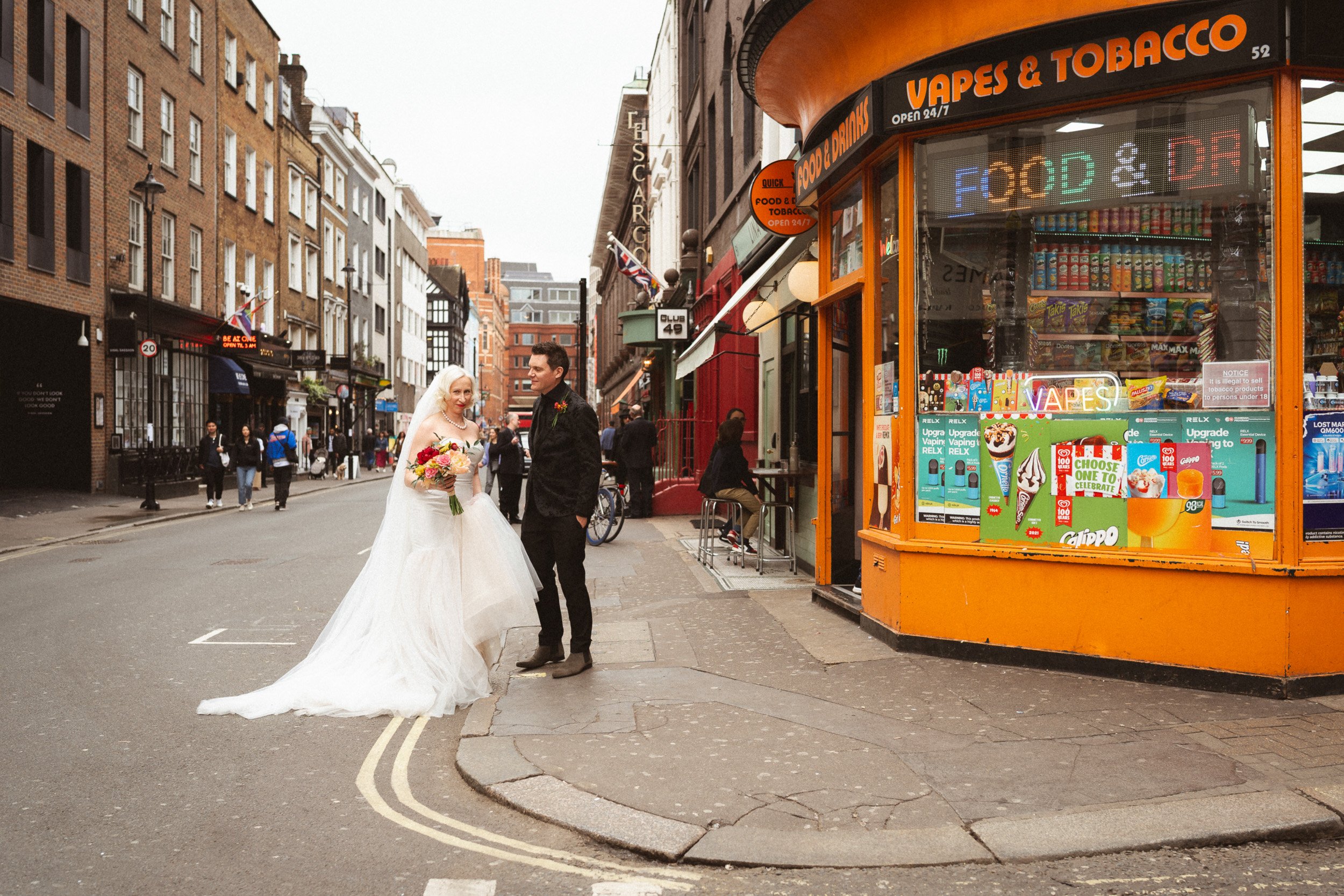 Bride and groom standing on a Soho street corner outside a newsagent near The Union Club after their Fitzrovia Chapel wedding in London