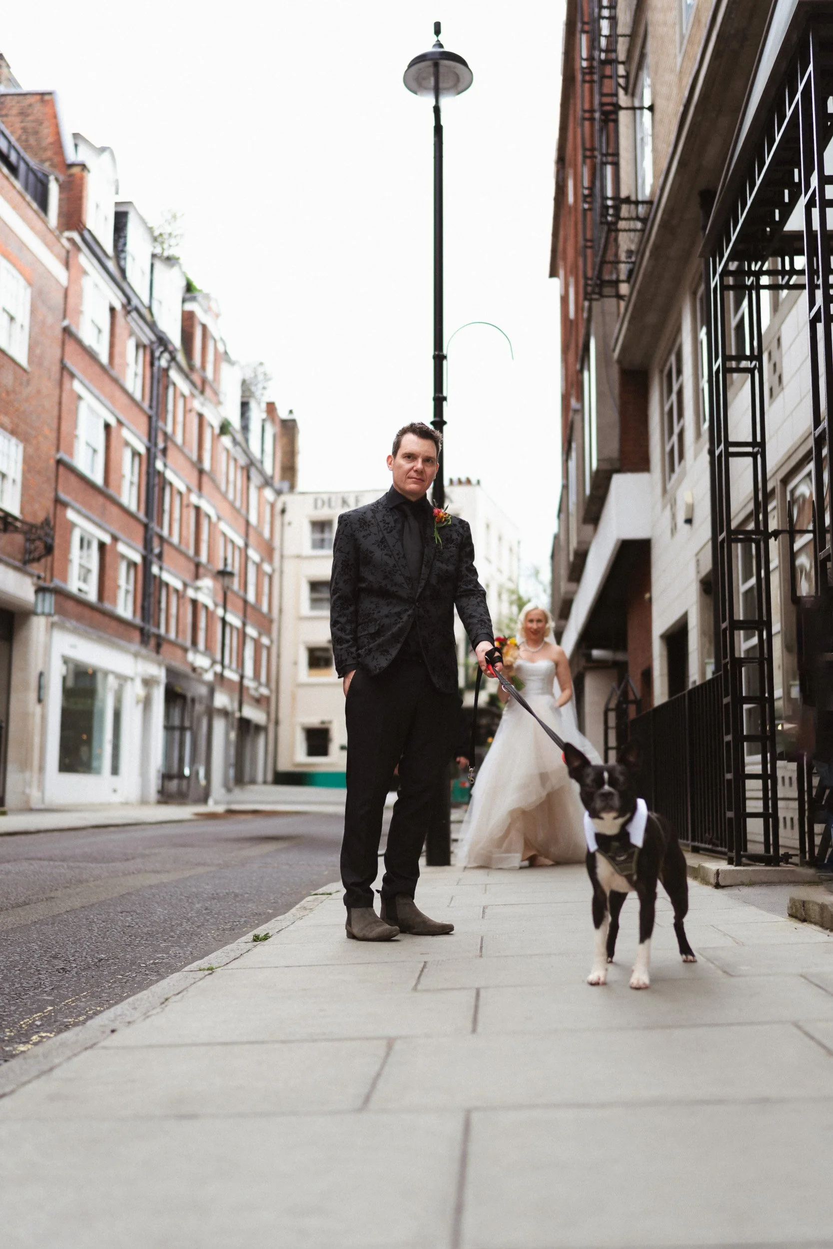 Groom standing on a Soho street  with his dog and the bride in the background - near The Union Club after their Fitzrovia Chapel wedding in London