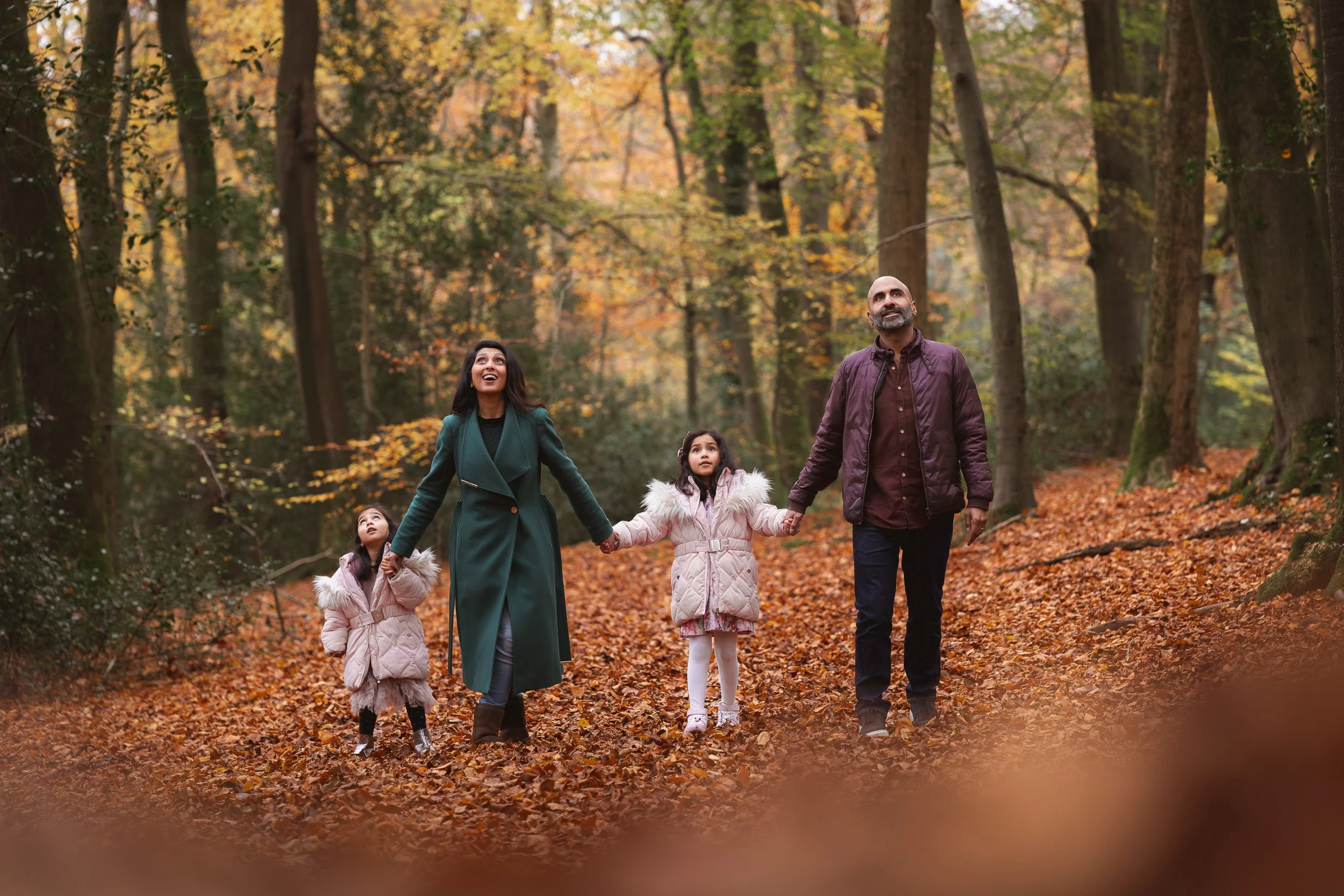 A young family of 4 walk  among  autumn leaves during a natural outdoor family photography session in the countryside near Thame, Oxfordshire