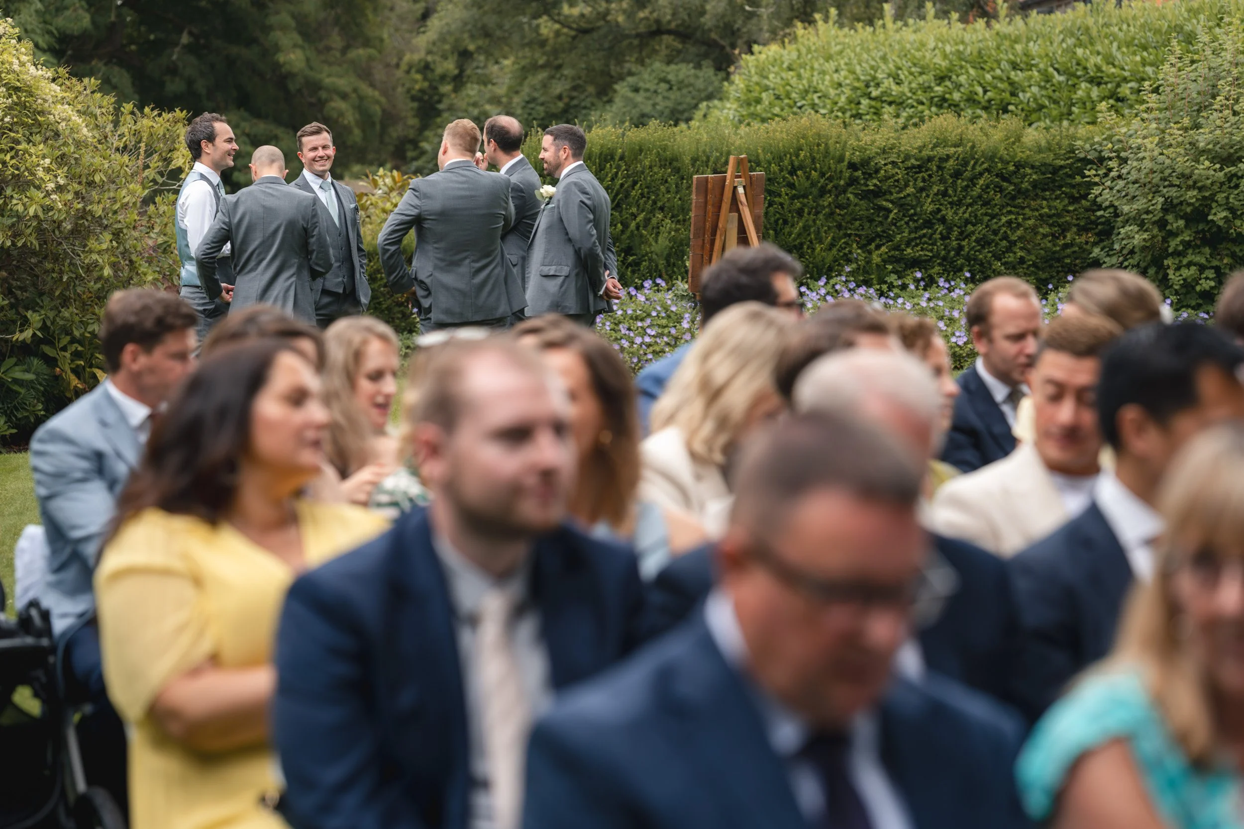 Wedding guests taking their seats with the Groom laughing with his groomsmen  on the lawn before an outdoor ceremony at Wasing Park in Berkshire