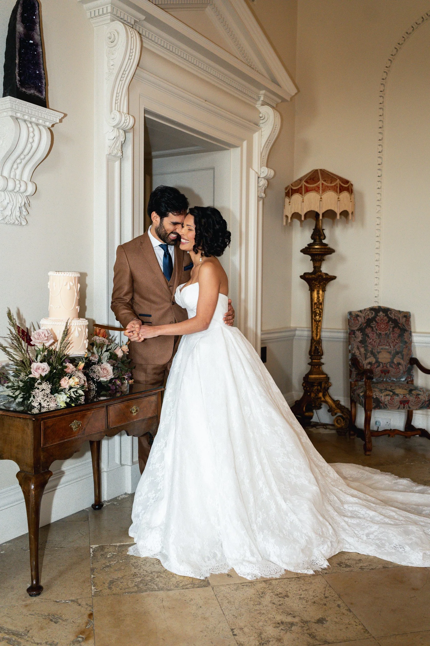 Bride and groom laughing and kissing as they cut their pink floral wedding cake on a flower lined table at their wedding reception at Kirtlington Park in Oxfordshire