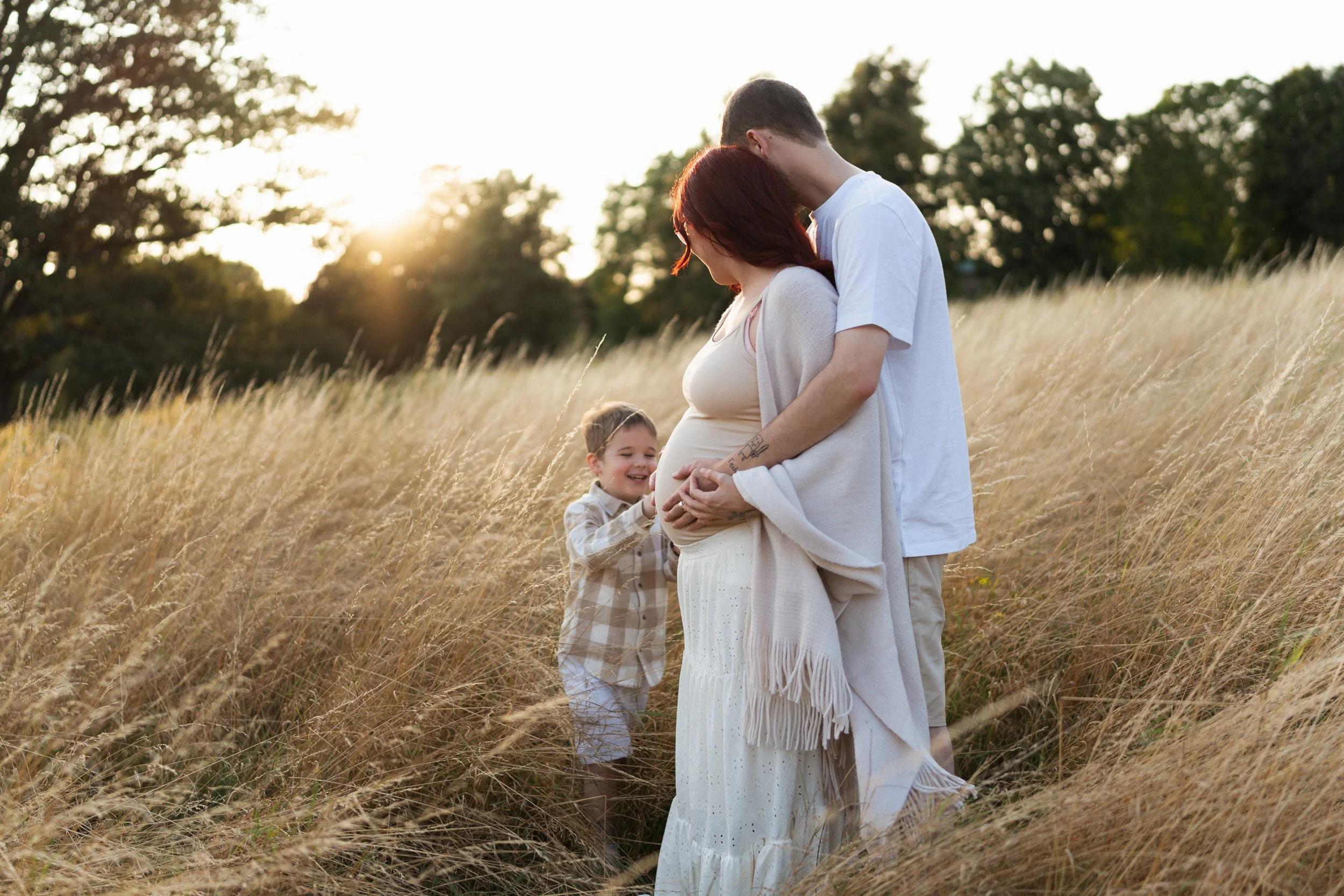 Husband embracing his pregnant wife at sunset while their young son gently touches her belly during a family session in Buckinghamshire