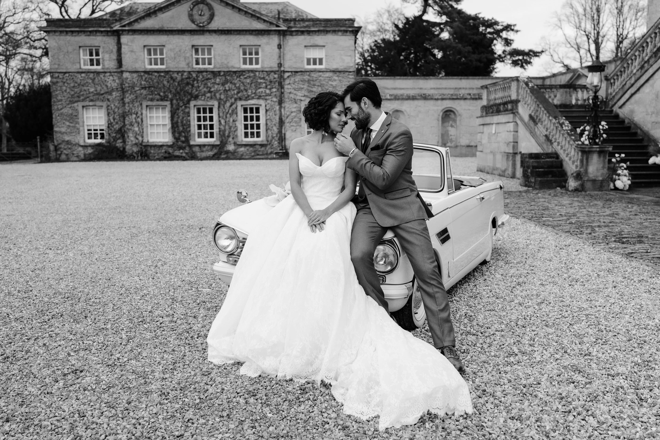 Bride and groom leaning in for a kiss on a vintage white wedding car at Kirtlington Park in Oxfordshire