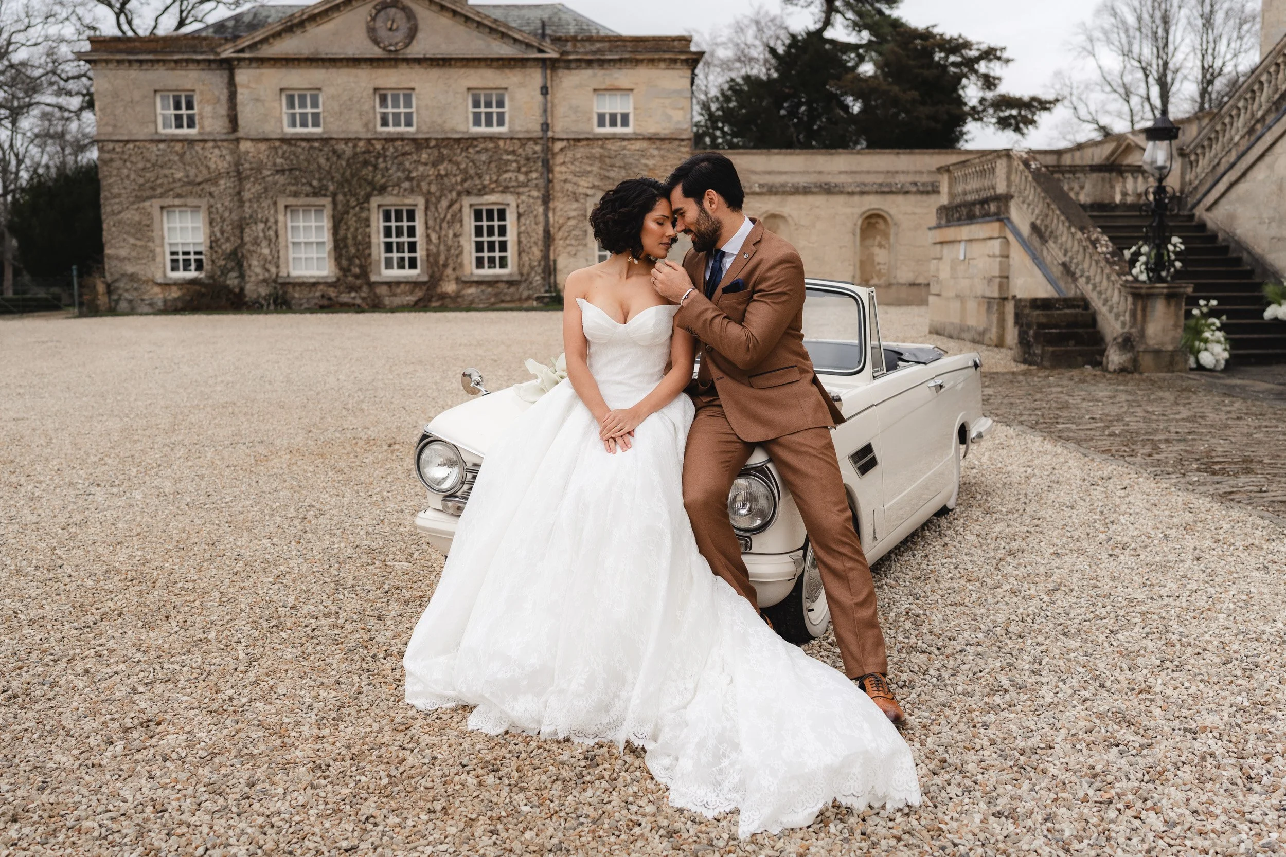 Bride and groom leaning in for a kiss on a vintage white wedding car at Kirtlington Park in Oxfordshire