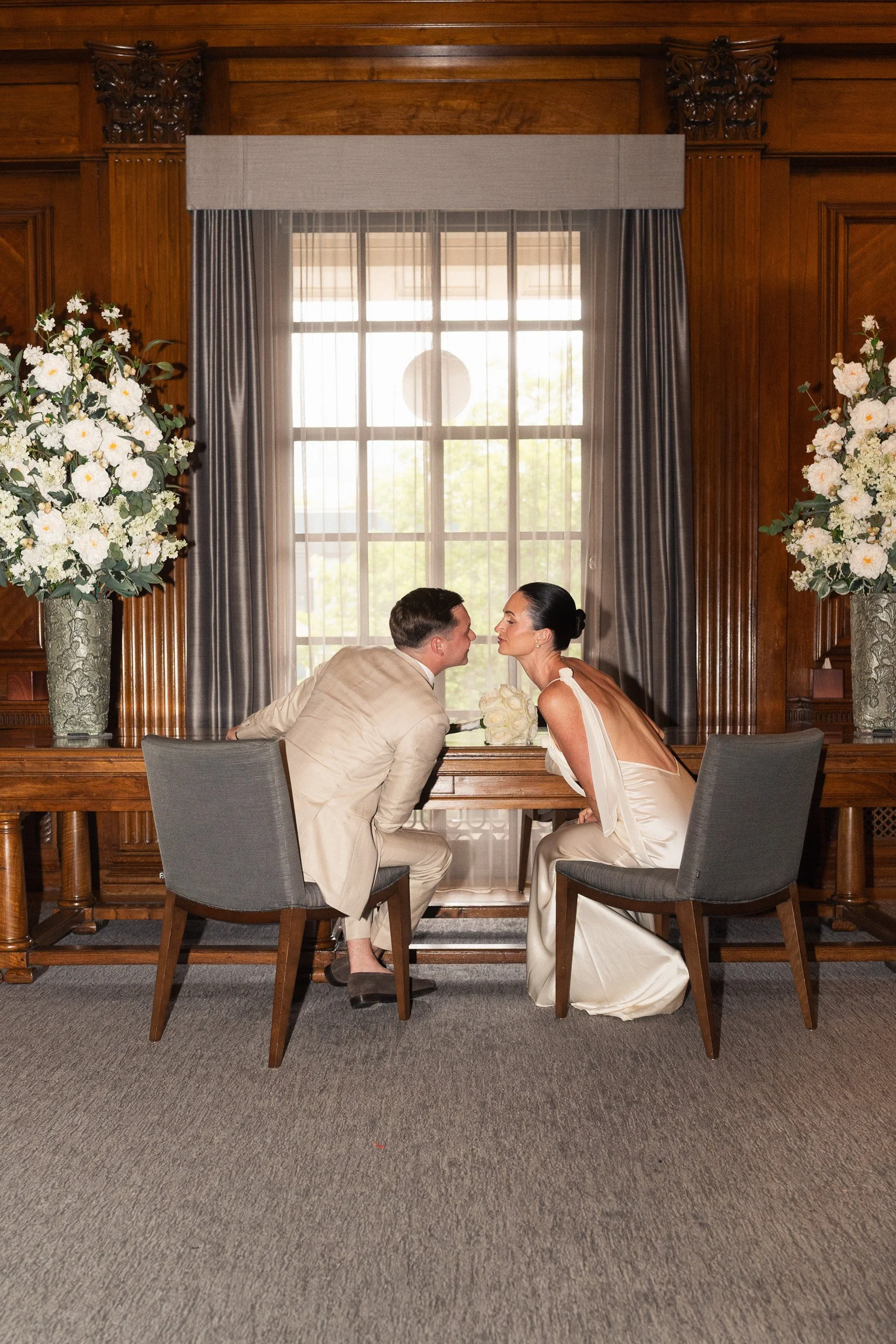 Bride and groom leaning across a table to kiss during their intimate wedding ceremony at Marylebone Town Hall Registry Office in London  