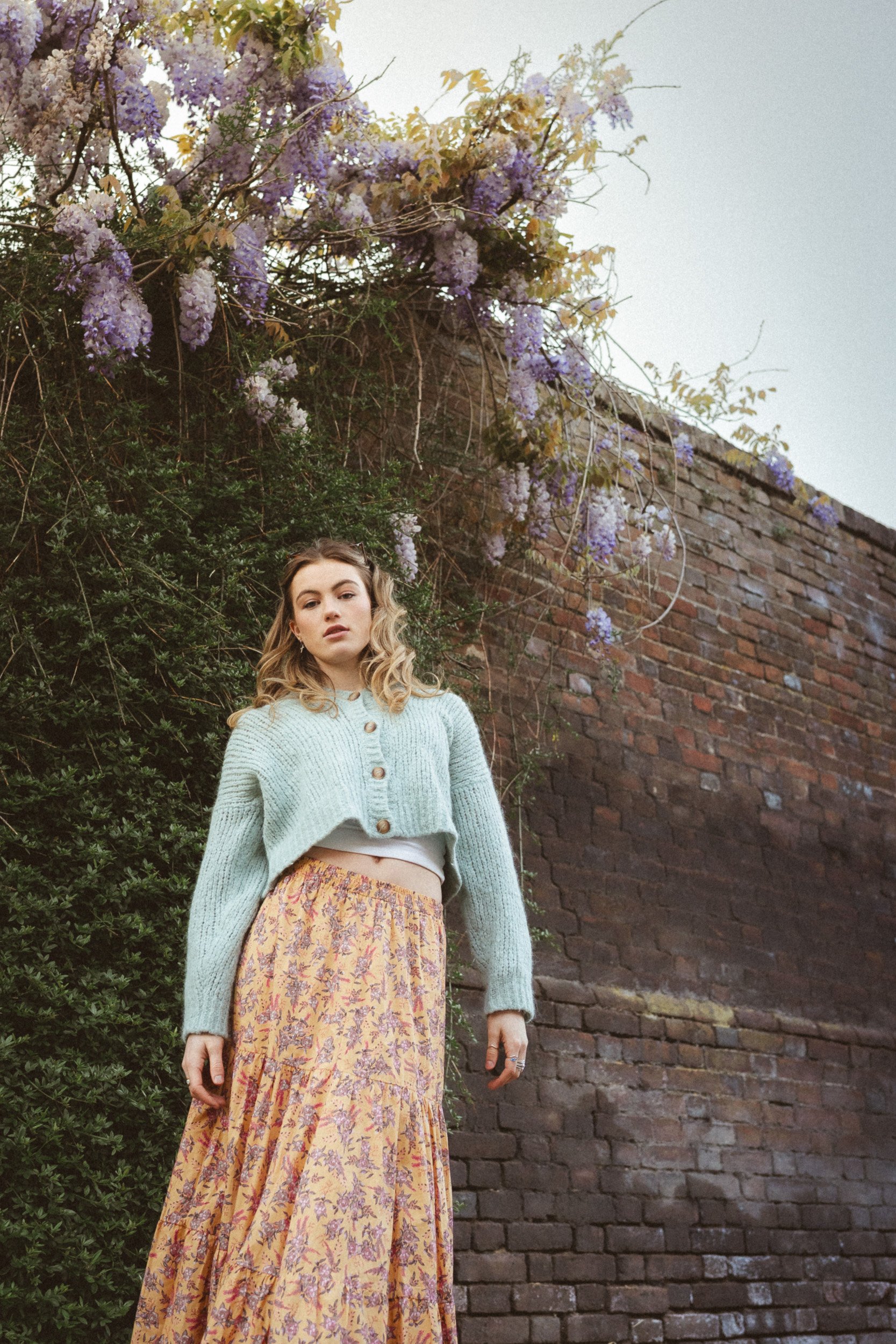 A young woman crossing the road with a garden wall covered with wisteria behind her during a school leaver photography session in Amersham, Buckinghamshire