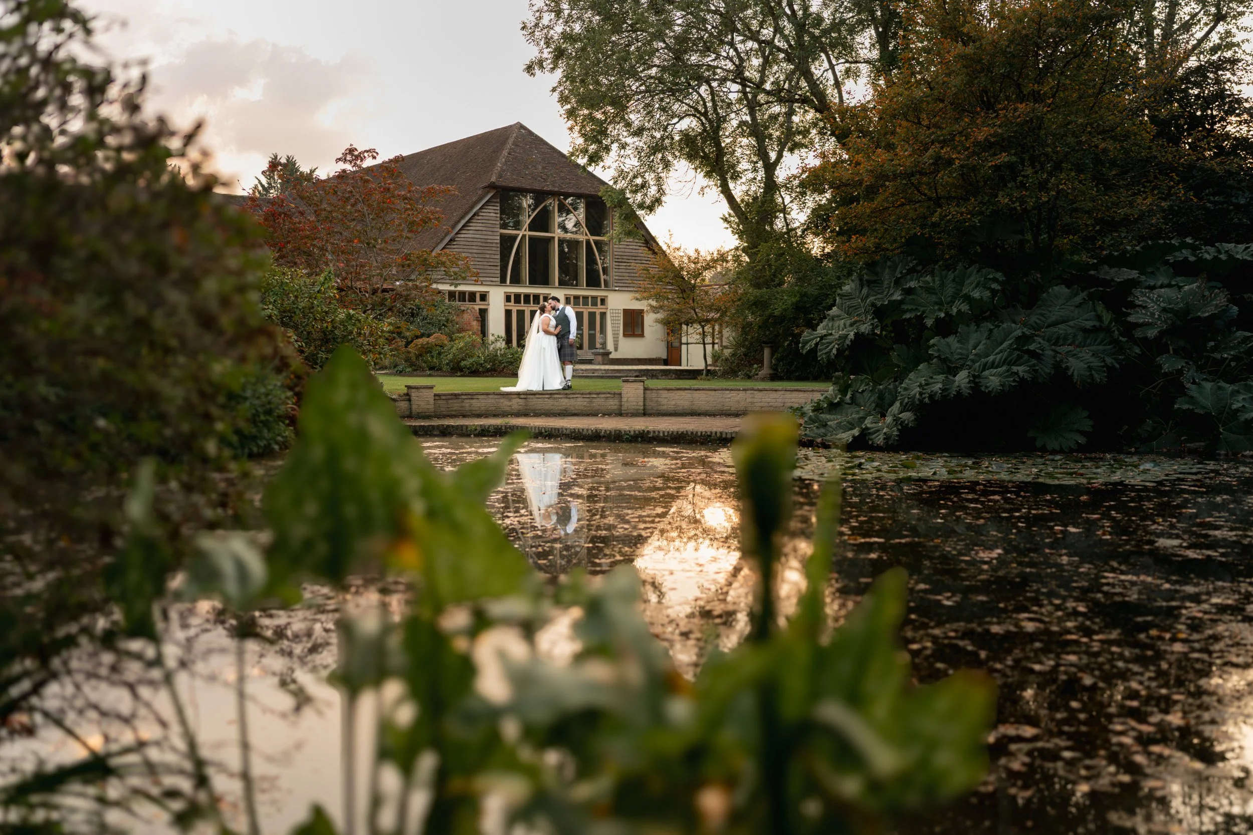 Bride and groom embracing in the golden hour light from across the pond at their Rivervale Barn wedding venue in Hampshire