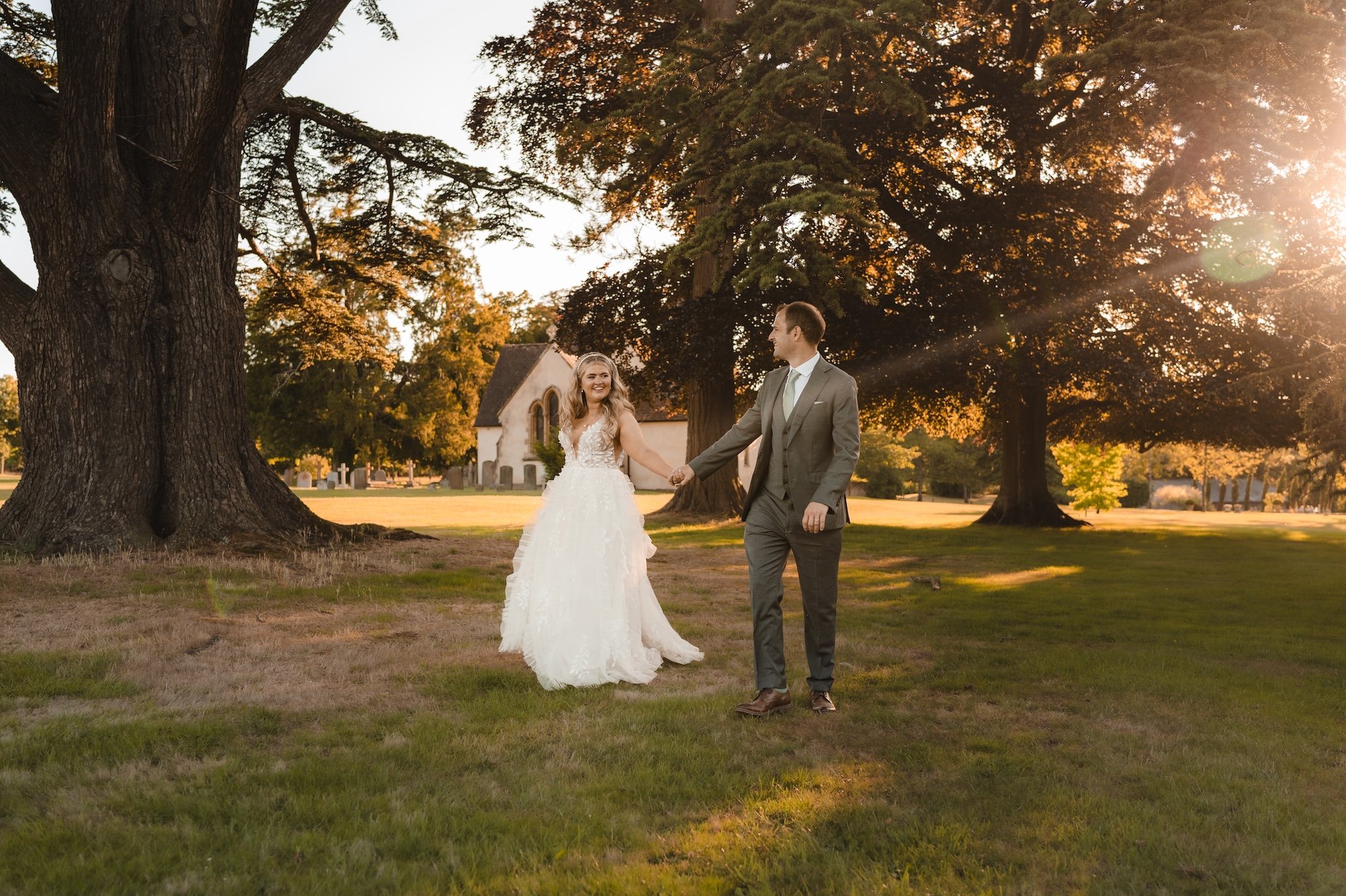 Bride and groom walking through Wasing Park at golden hour with long evening shadows and St Nicholas Church behind them