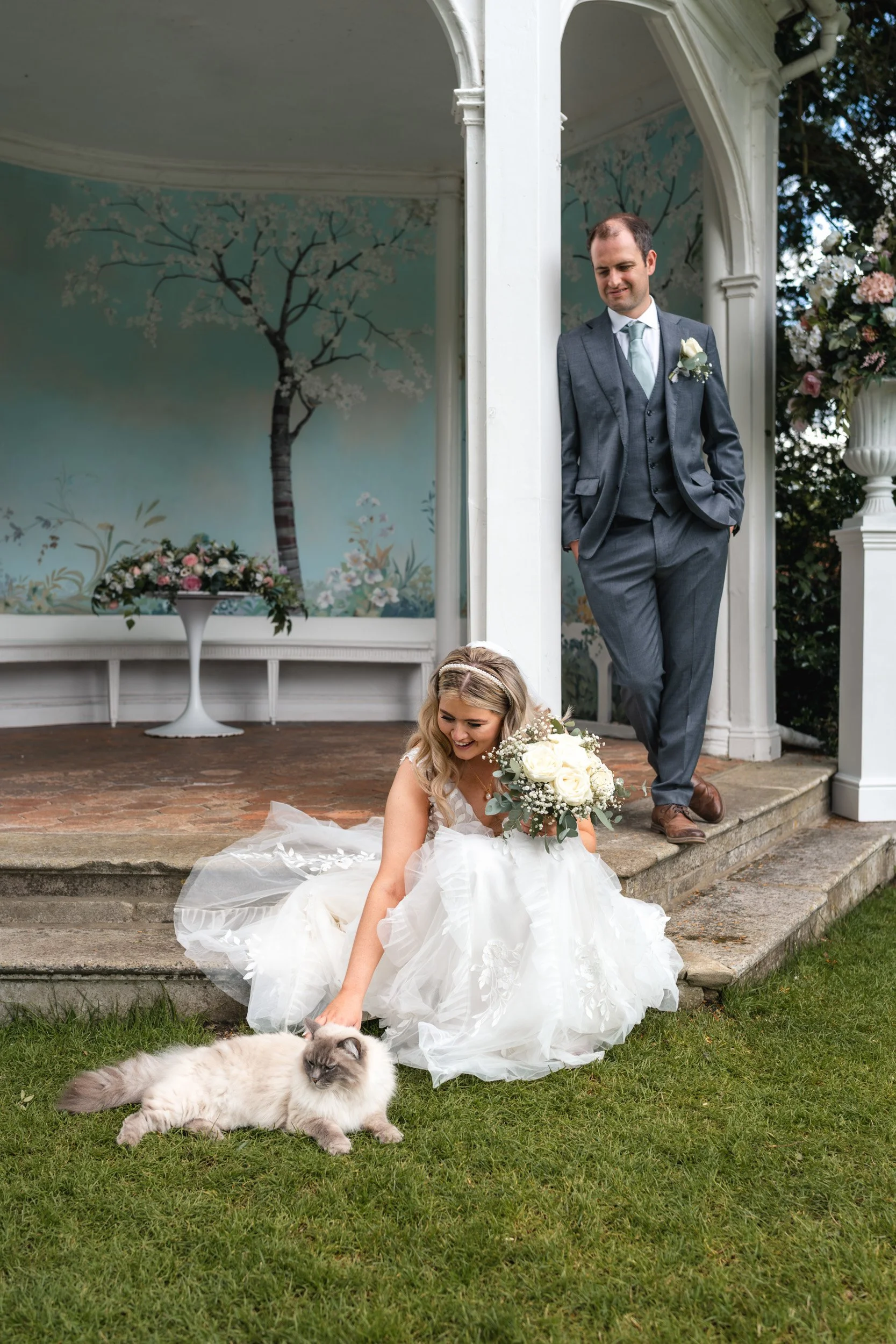 Bride crouching to stroke the  resident cat on the lawn Wasing Park  in Berkshire while the groom stands behind looking happy