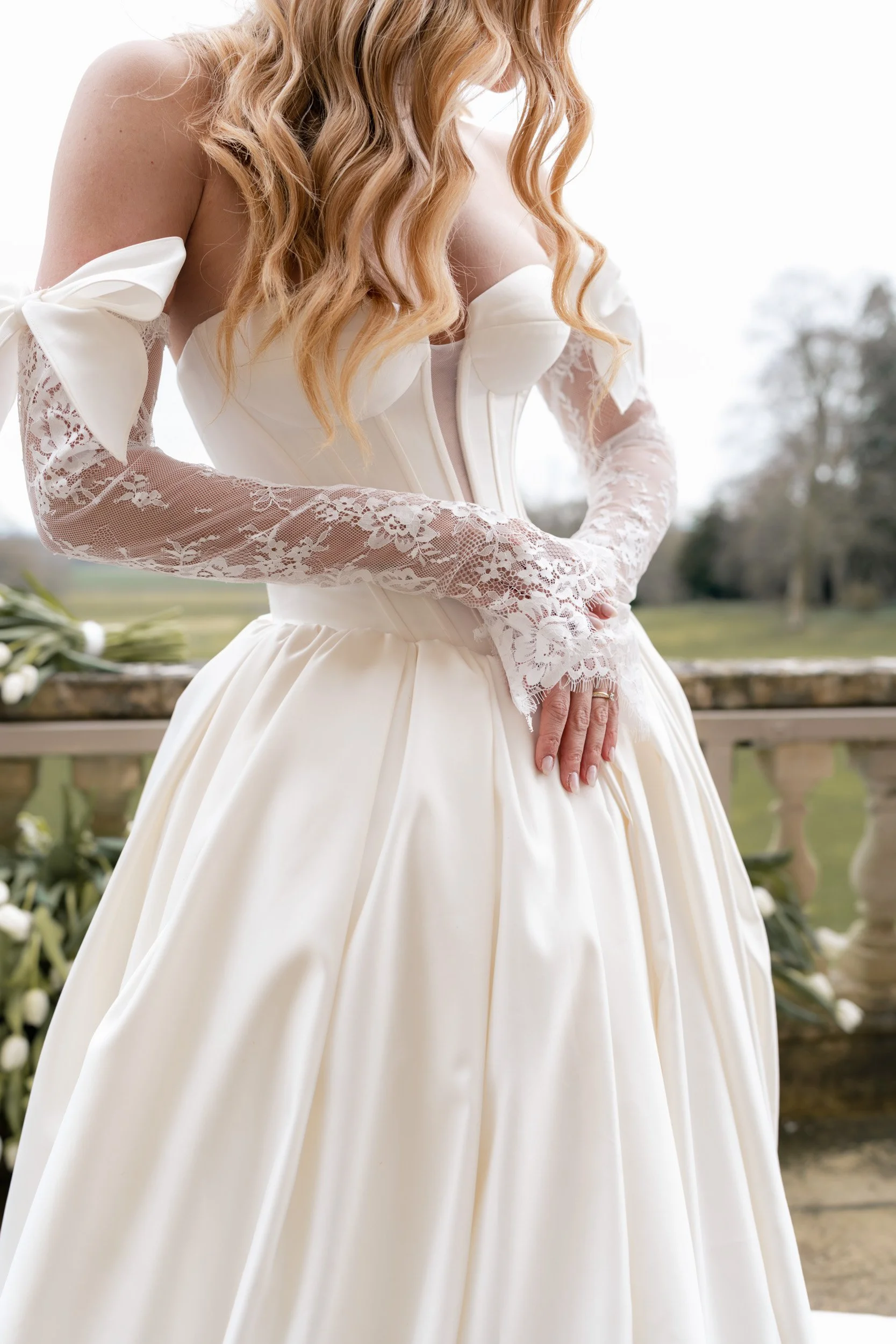close up of a bride looking down at her dress and fixing her lace gloves on the flower lined balcony over looking the garden and grounds of Kirtlington Park in Oxfordshire