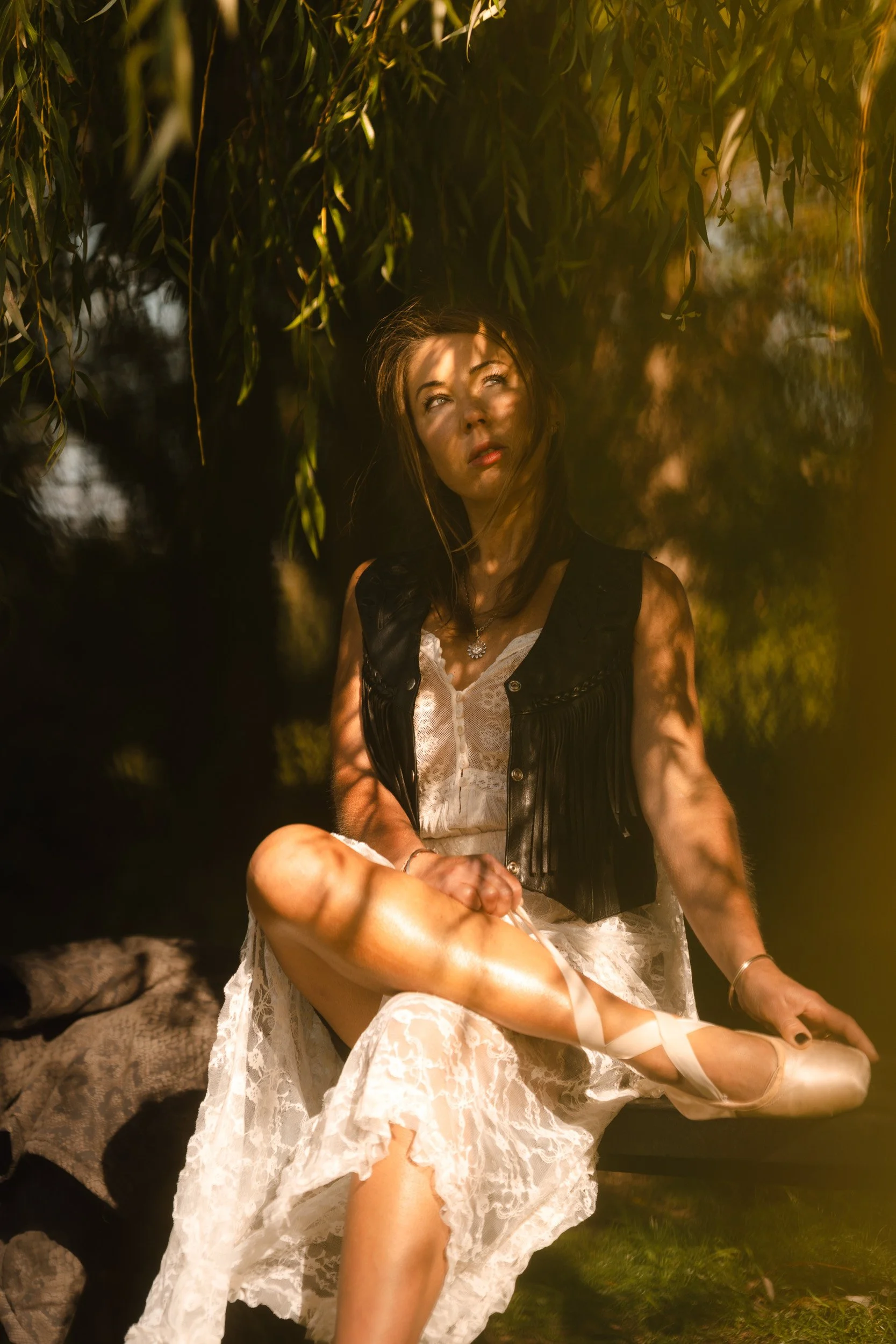 Woman sitting beneath a willow tree in dappled sunlight in Windsor, Berkshire wearing a leather waste coat and ballet slippers