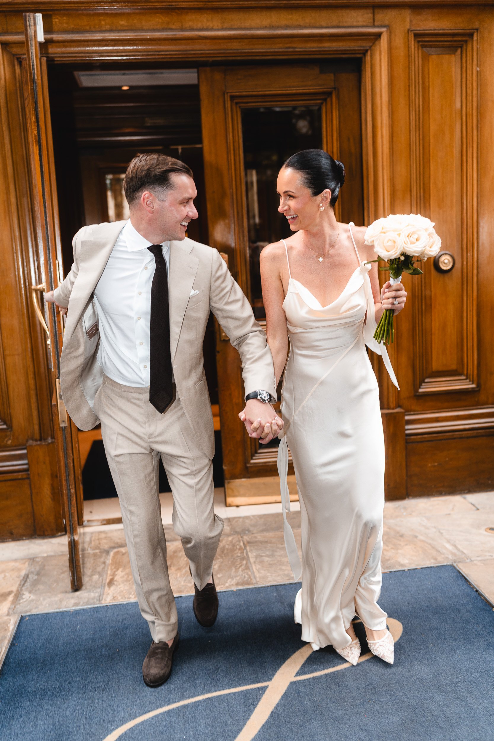 Bride and groom leaving The Landmark Hotel hand in hand and laughing to head to their wedding ceremony at Marylebone Registry Office in London