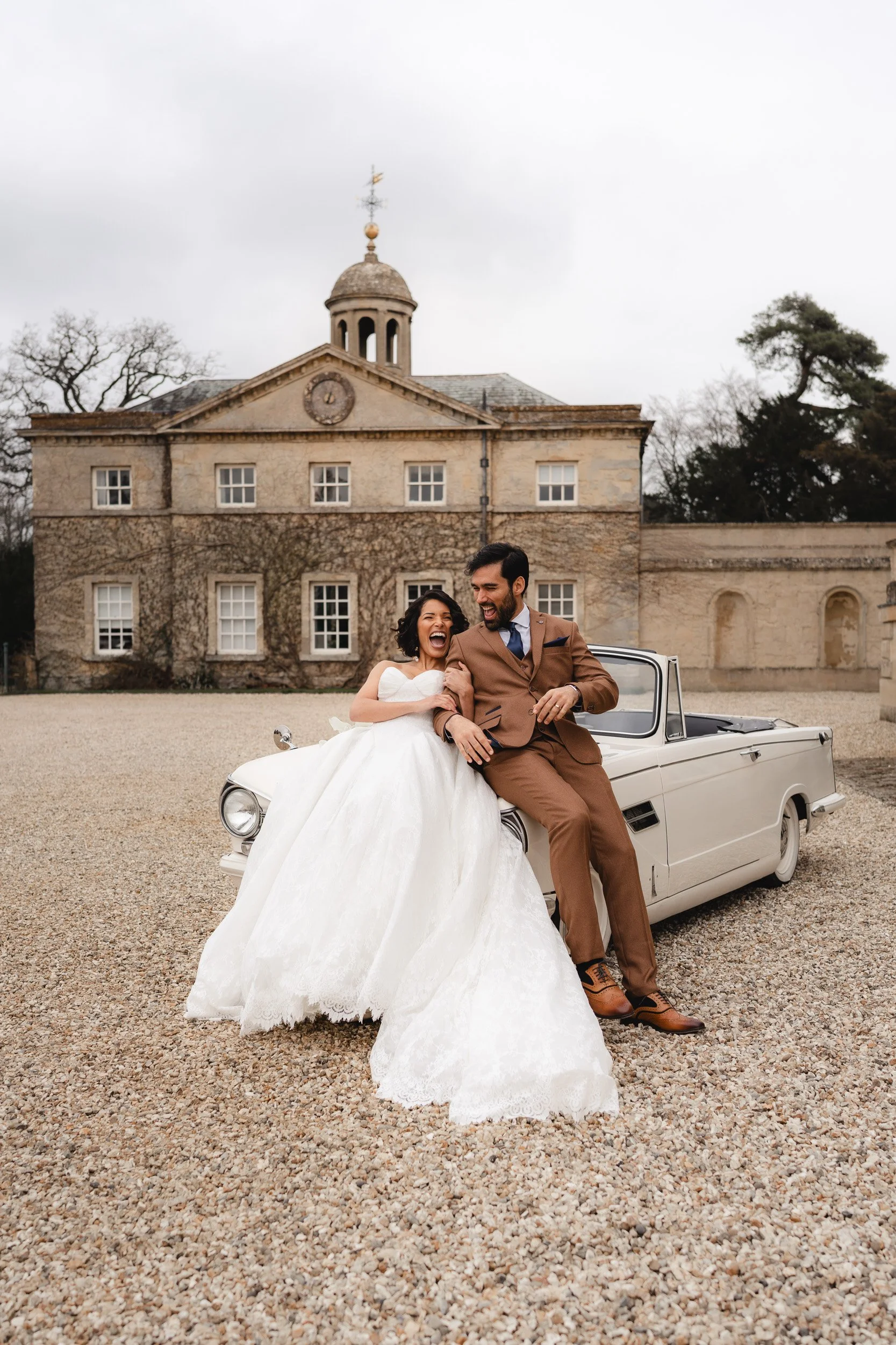 Bride and groom laughing while leaning on a vintage white wedding car at Kirtlington Park in Oxfordshire