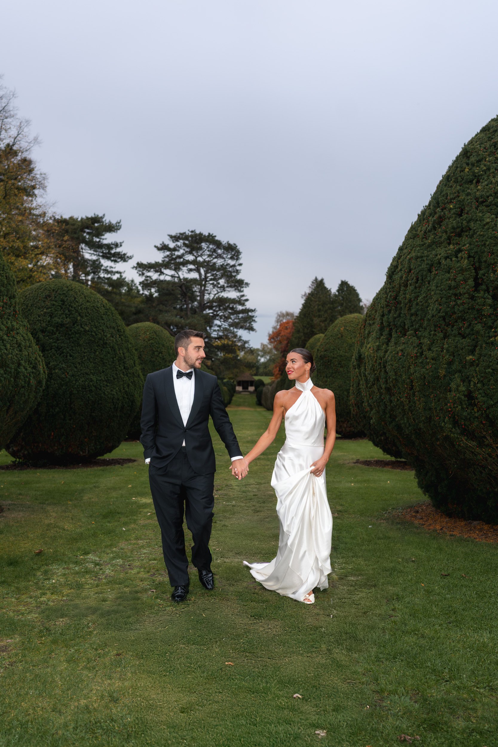 Bide and groom walking hand in hand through the tree lined gardens and lawn of The Elvetham Hotel in Hampshire