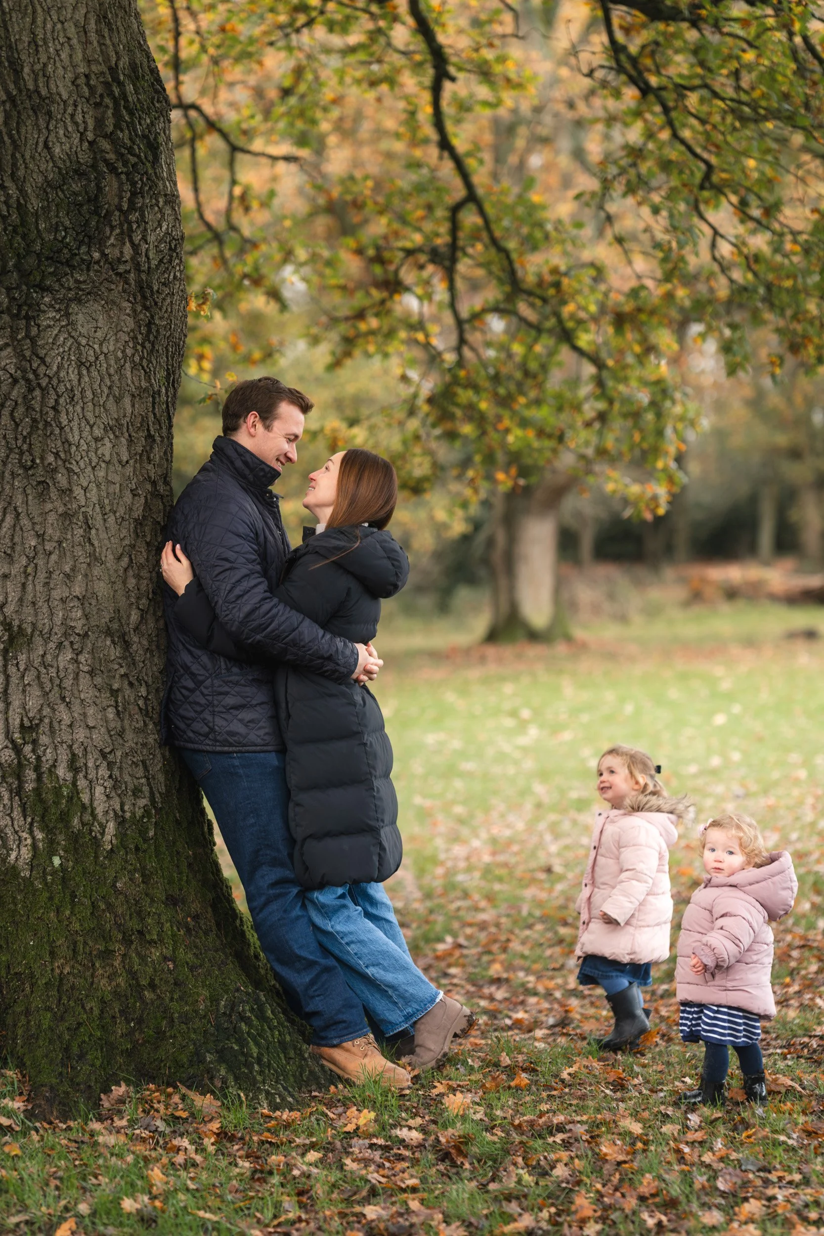 Two little girls look up to the parents having a cuddle under an oak tree during a family photography session in Amersham, Buckinghamshire