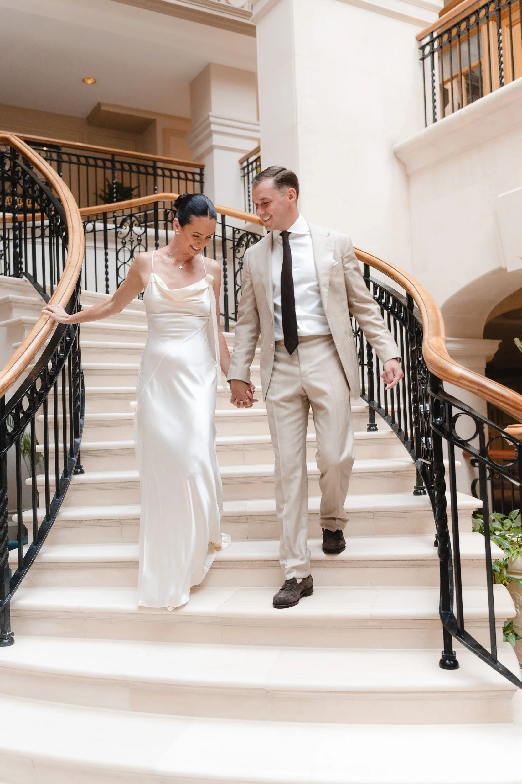 Bride and groom walking down the grand staircase hand in hand and laughing at The Landmark Hotel,  London in Marylebone