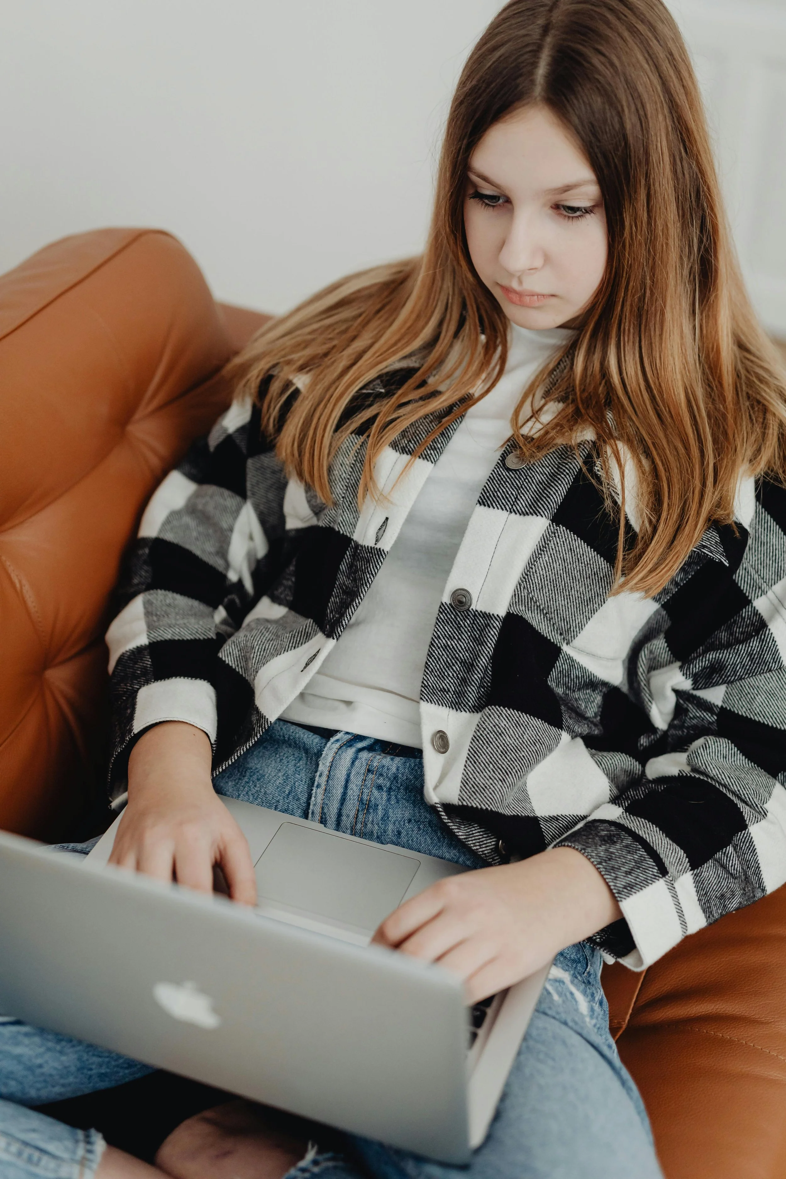A young woman with red hair wearing a black and white plaid shirt and jeans, sitting on a brown leather couch, using a silver MacBook laptop.