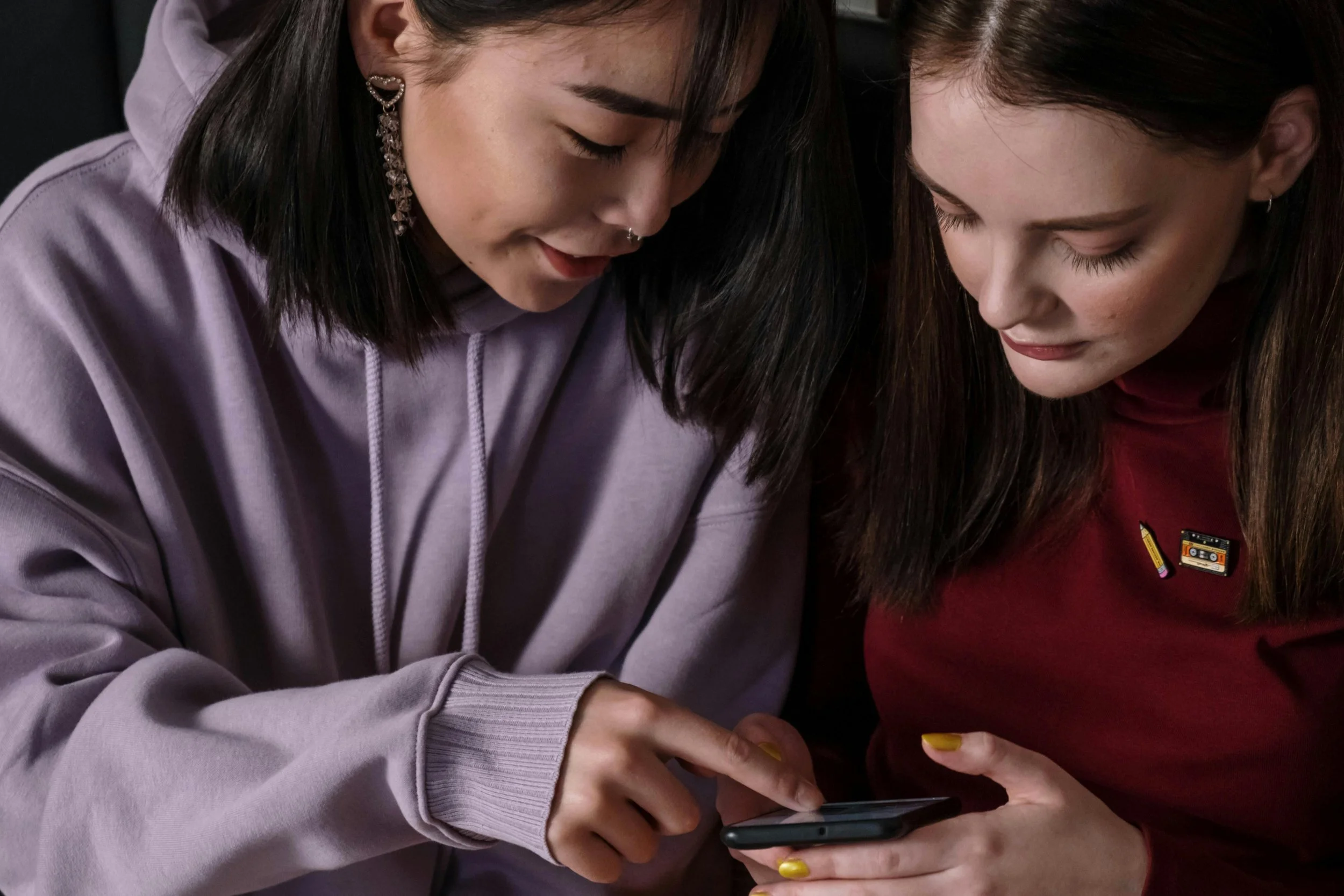 Two young women looking at a smartphone together, one pointing at the screen, both with focused expressions.