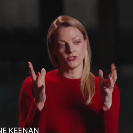 A woman with red hair wearing a red top, speaking and gesturing with her hands.