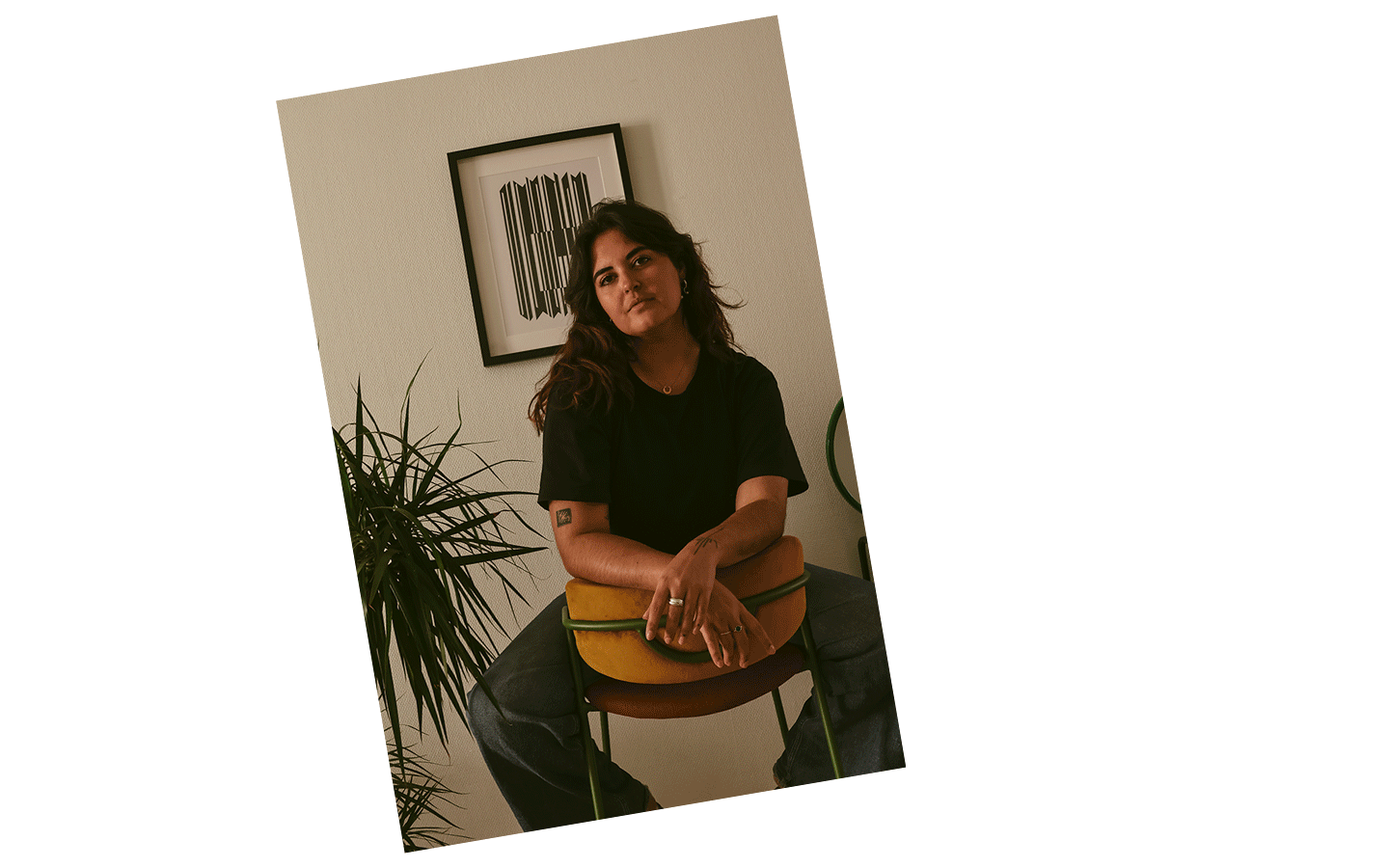 Une jeune femme brune assise sur une chaise haute orange et verte, portant un t-shirt noir et un jean bleu. Il y a une plante verte à gauche et un tableau de Vasarely avec un motif géométrique en noir et blanc en arrière-plan.