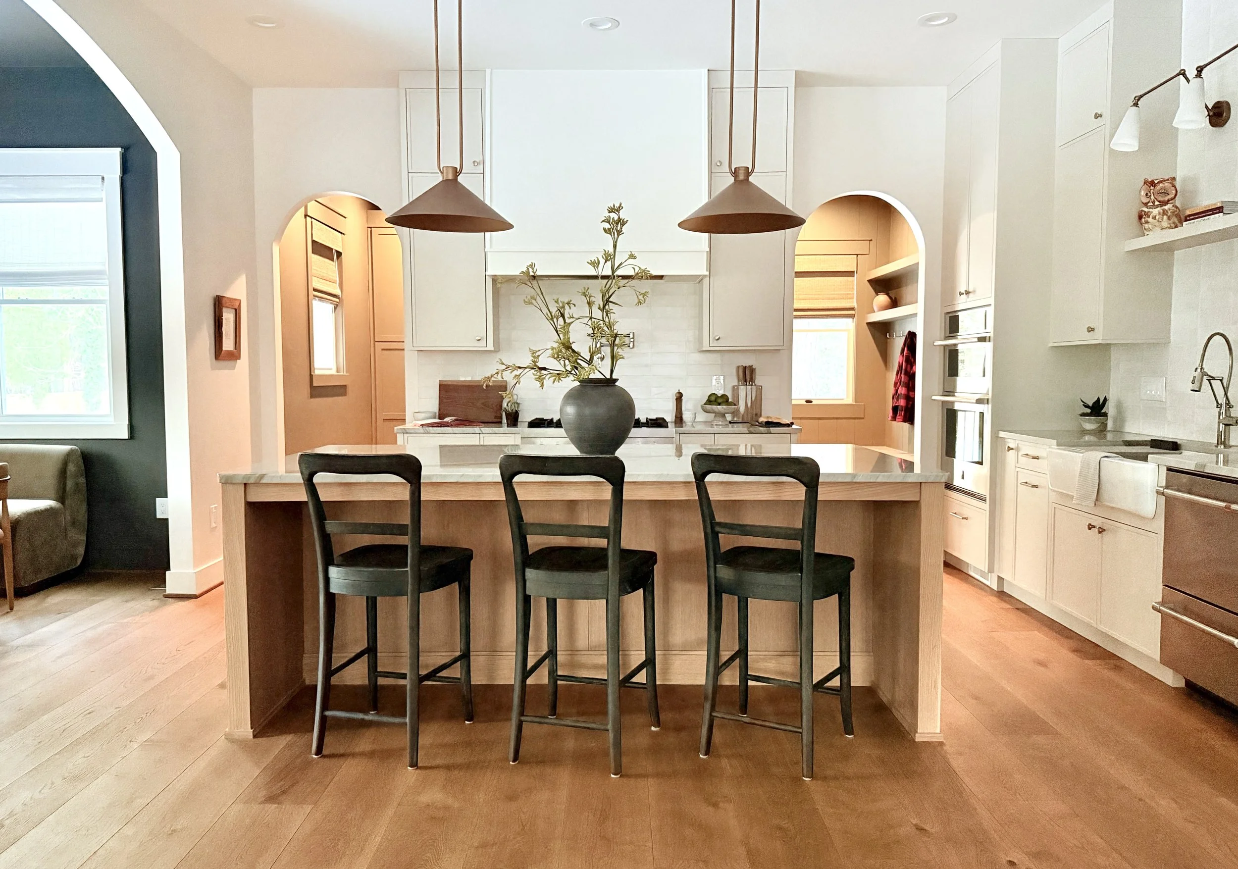 Modern kitchen with a large island, black chairs, white cabinets, and pendant lights, with a window and decorative items.
