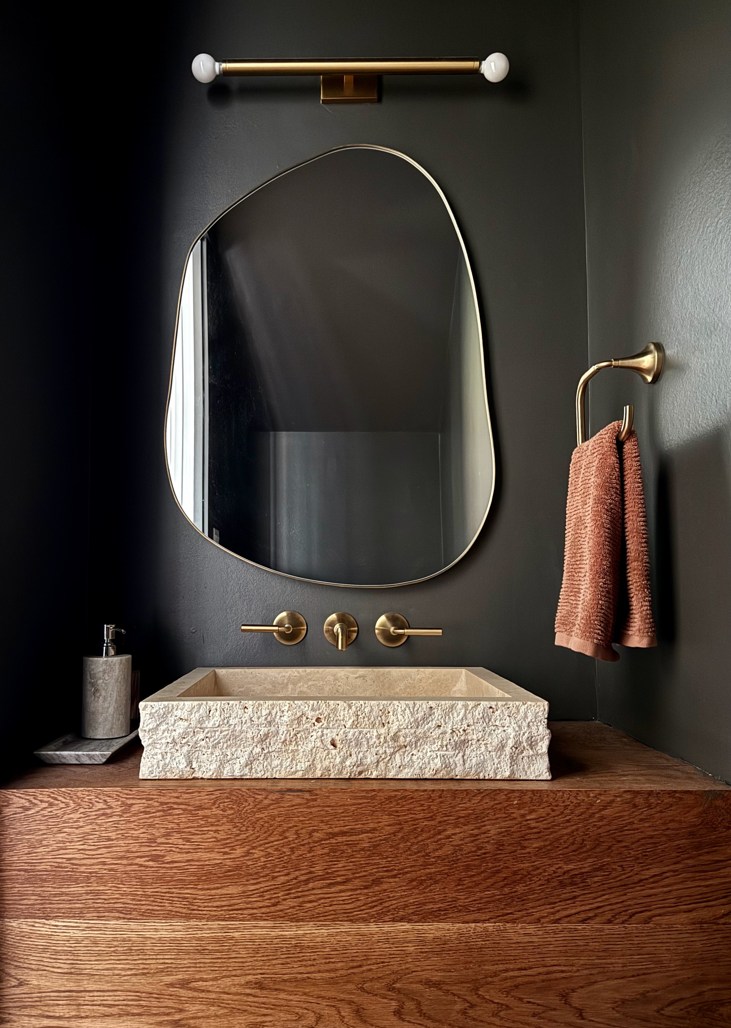 Powder room featuring a textured travertine vessel sink and dark taupe gray walls. Brass wall mounted fixtures, a sculptural organic mirror, and a white oak floating vanity create a warm, sophisticated aesthetic.