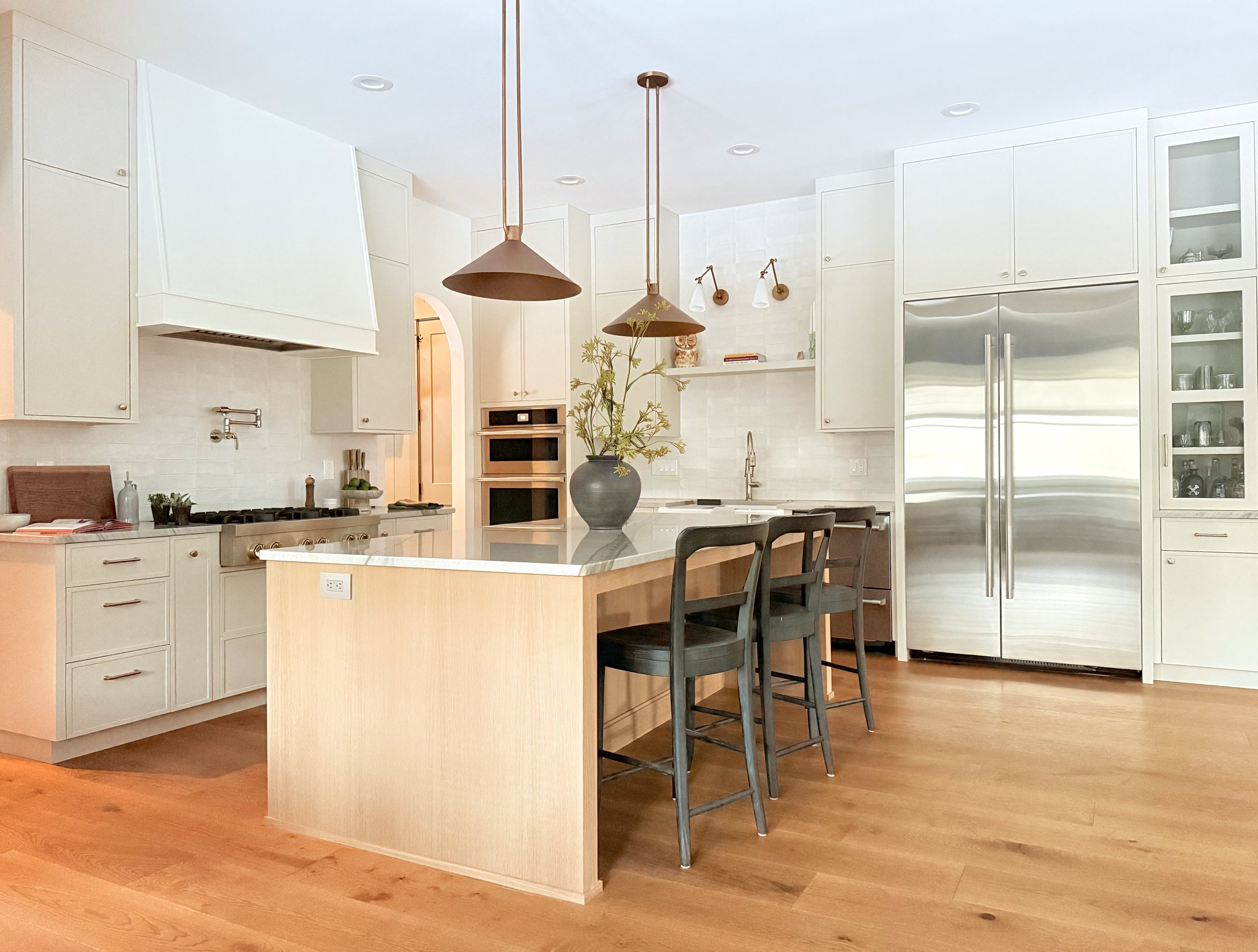 Modern kitchen with white cabinets, stainless steel fridge, wooden flooring, black chairs at a kitchen island, with pendant lights and a vase with green plants on the island.